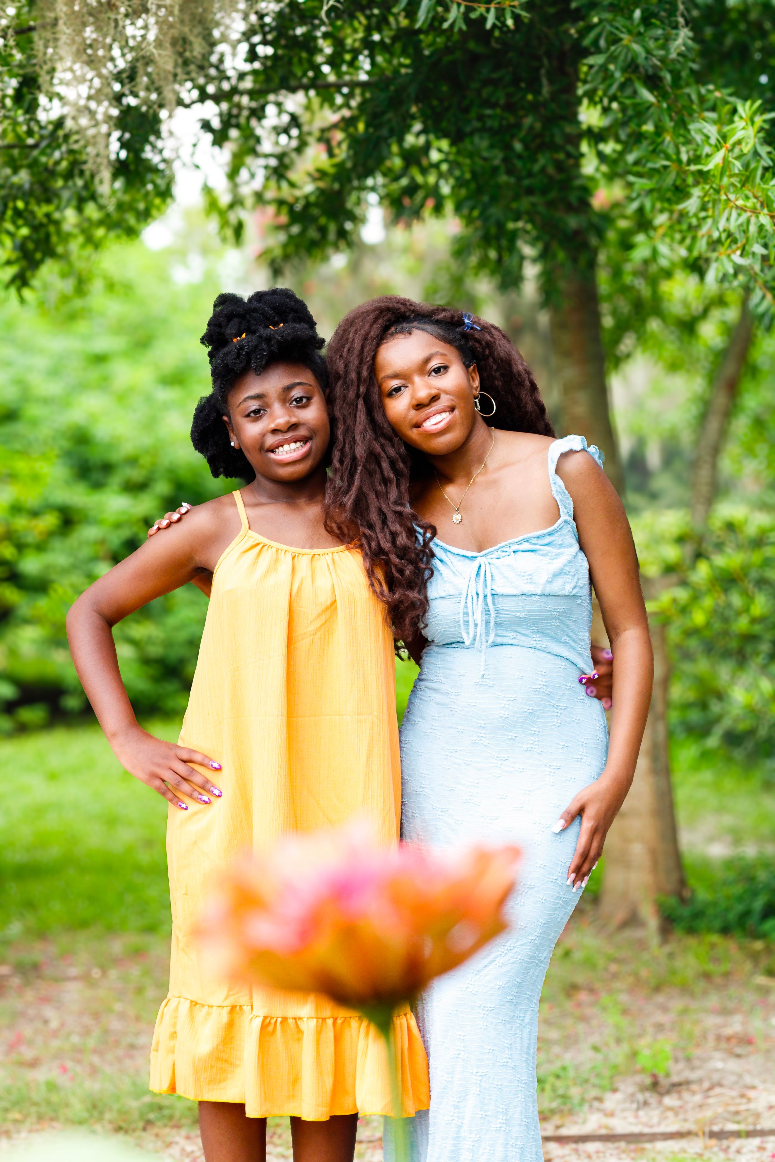 Two young Black women smiling outdoors in a green park, wearing yellow and blue dresses, with flowers in foreground.