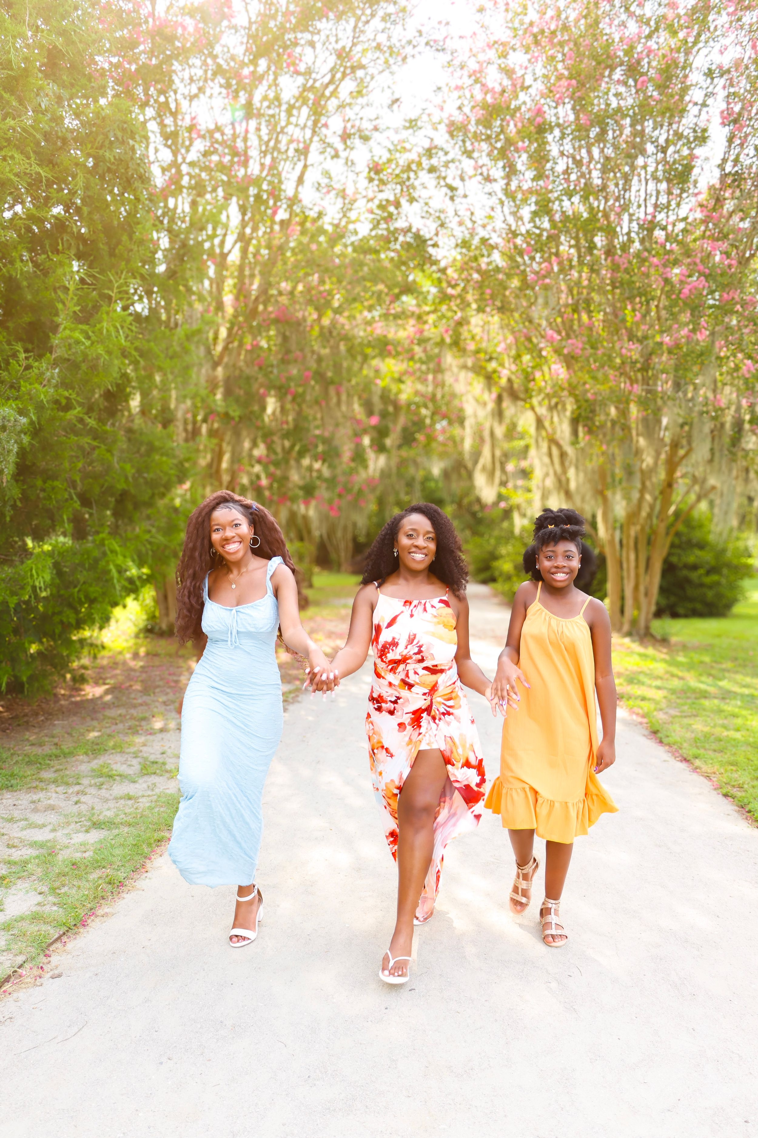 Three joyful women in colorful summer dresses walk hand-in-hand down a tree-lined path with blooming pink flowers.