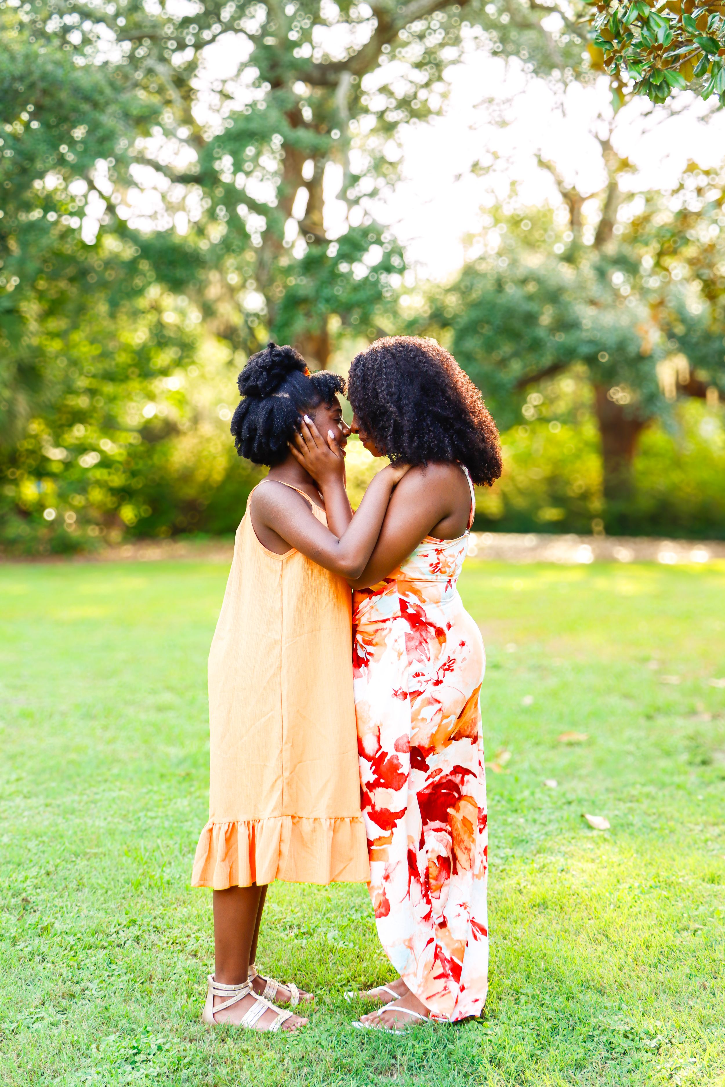 Two Black women in colorful summer dresses share a tender moment outdoors in a lush green park.