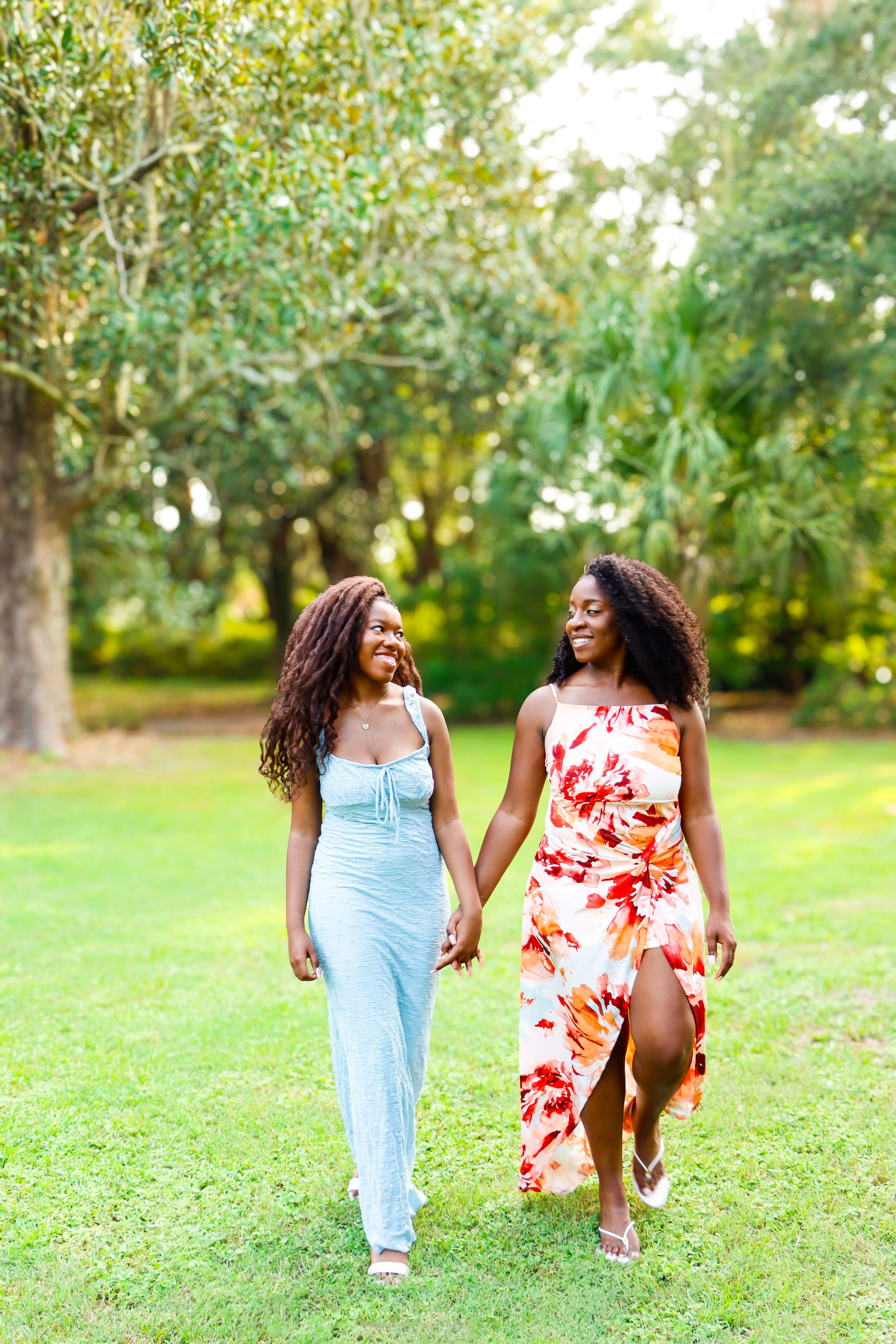 Two women in summer dresses hold hands while walking through a lush green park, smiling joyfully.