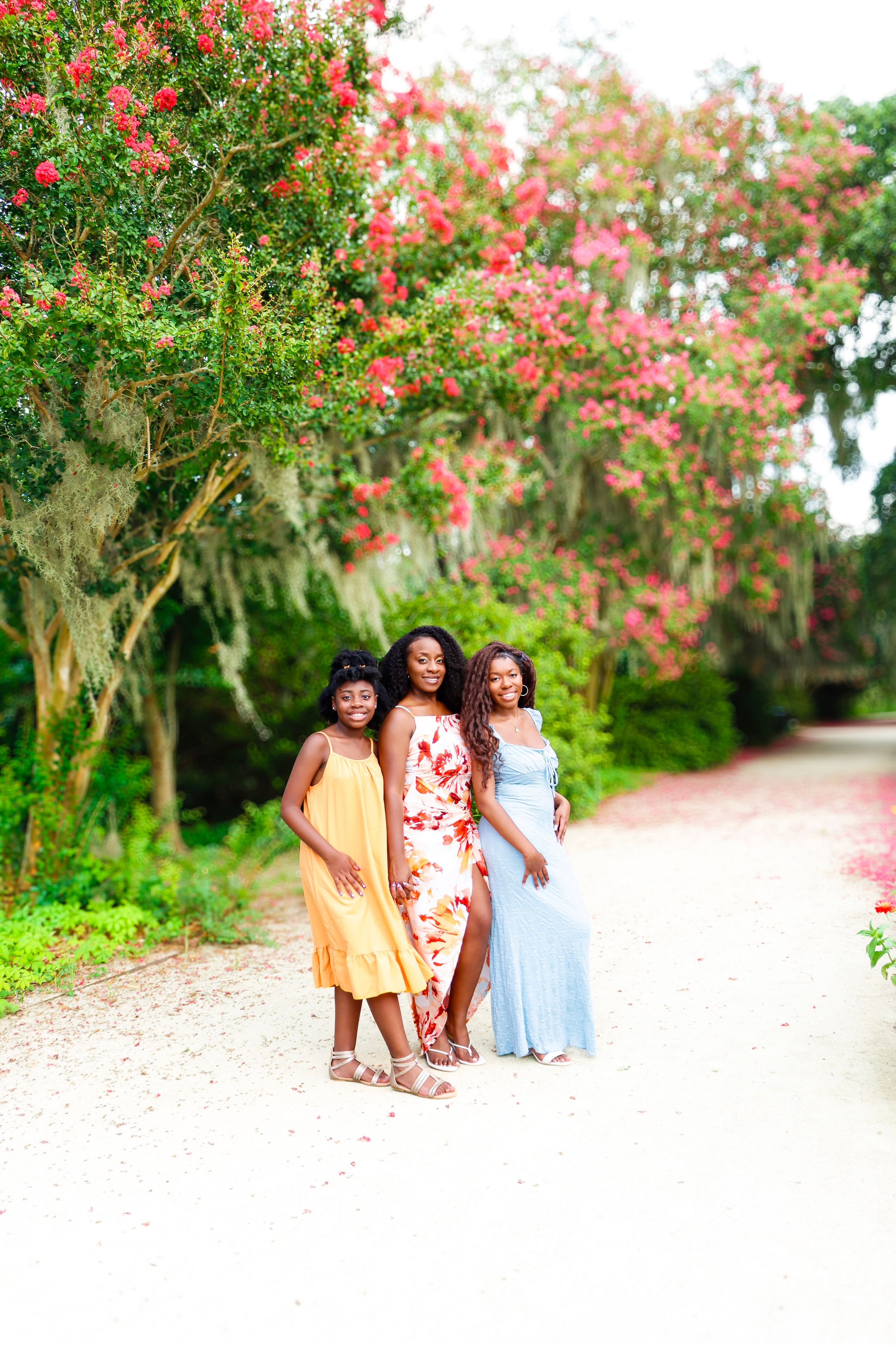 Three women in colorful summer dresses pose on a sandy path surrounded by lush green trees with pink blooming flowers.