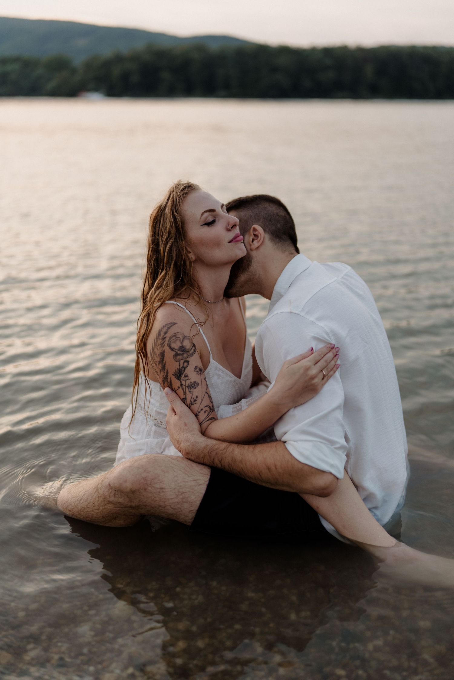A tattooed couple shares a romantic kiss while sitting in shallow lake water at sunset, dressed in white.
