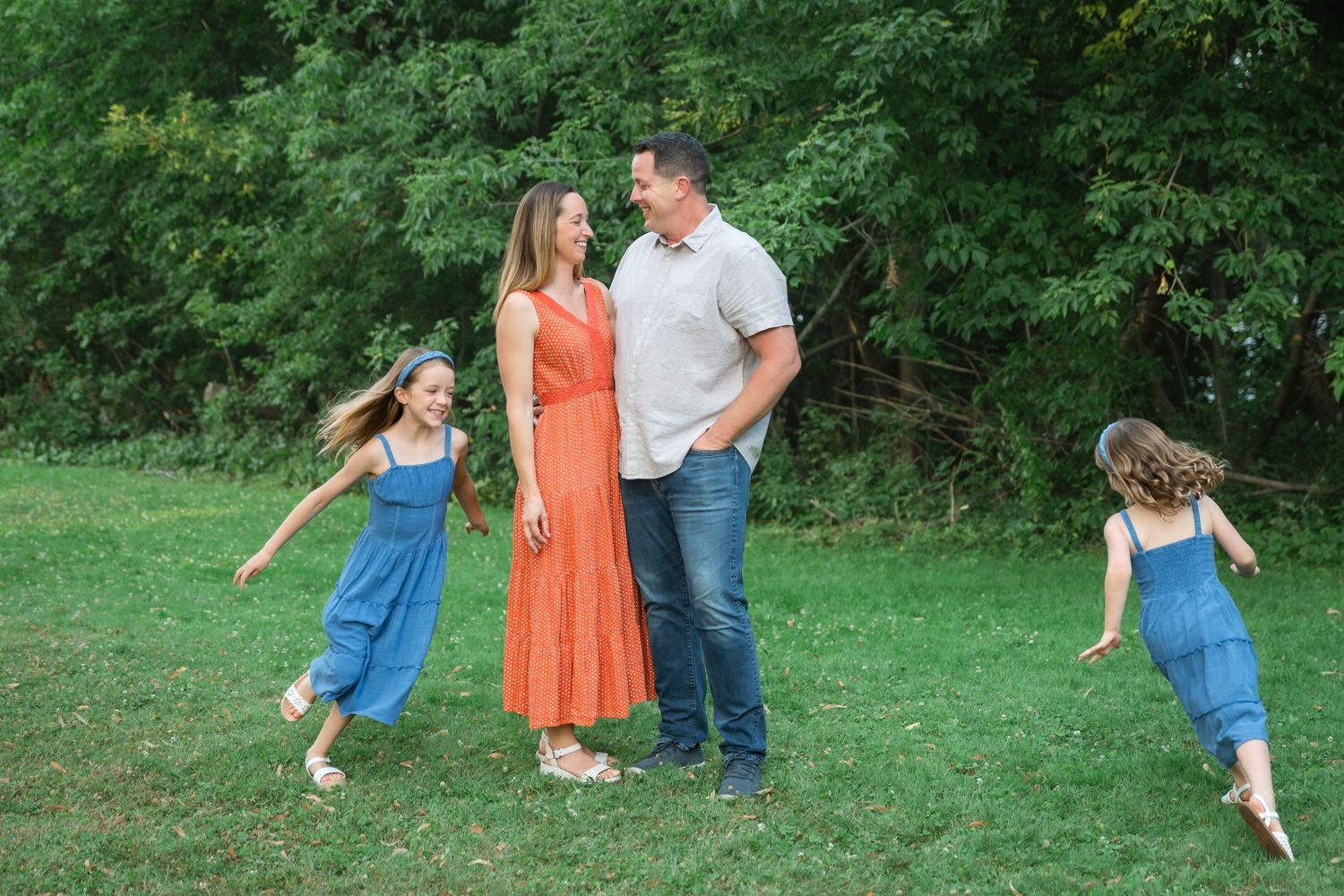 Family of four enjoying outdoor time on green grass, two girls in blue denim dresses spinning around parents in summer.