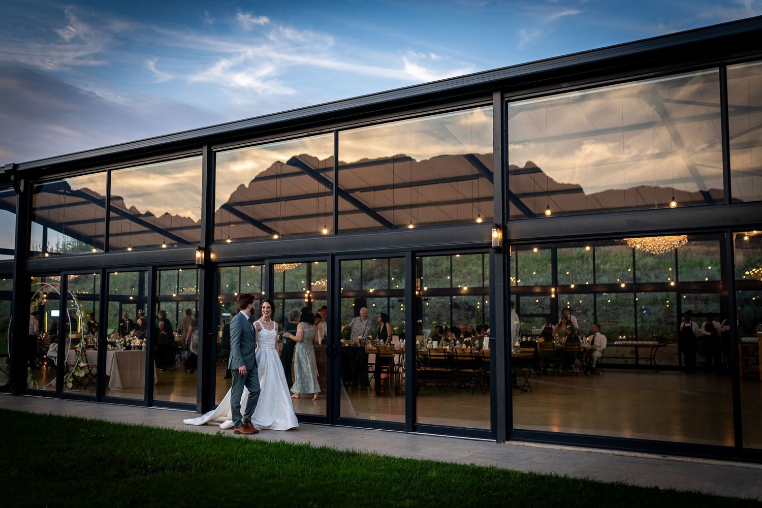 Bride and groom share a romantic moment outside an elegant glass-walled wedding venue at dusk.
