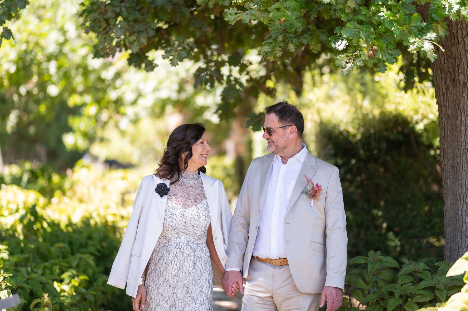 Couple in wedding attire holding hands in a lush garden, bride in lace dress, groom in tan suit with floral boutonniere.