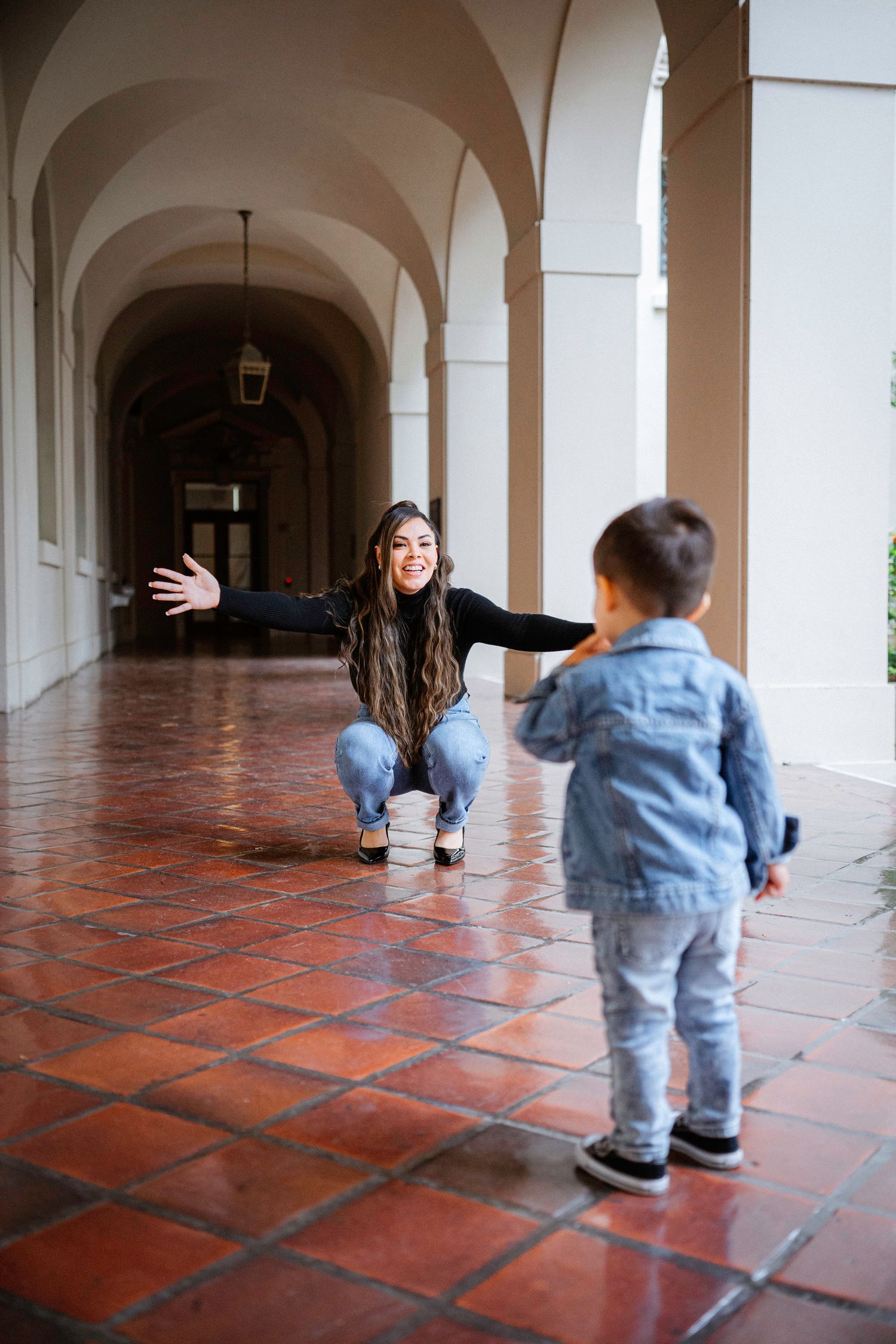 Woman crouching with open arms in arched hallway of Pasadena City Hall, welcoming a young child in denim jacket running toward her.