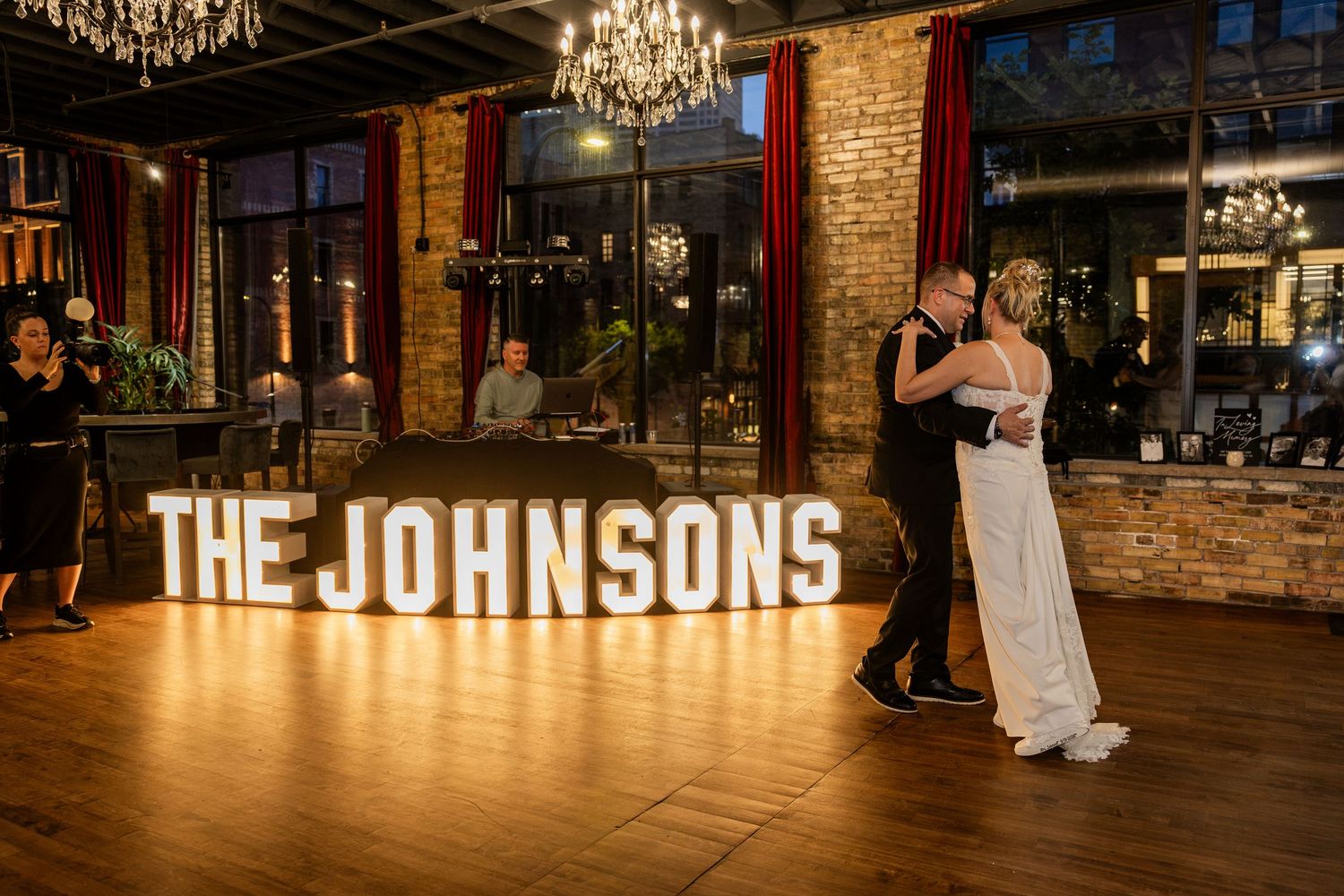 Newlyweds share first dance in front of illuminated THE JOHNSONS marquee letters at elegant brick-walled wedding reception.