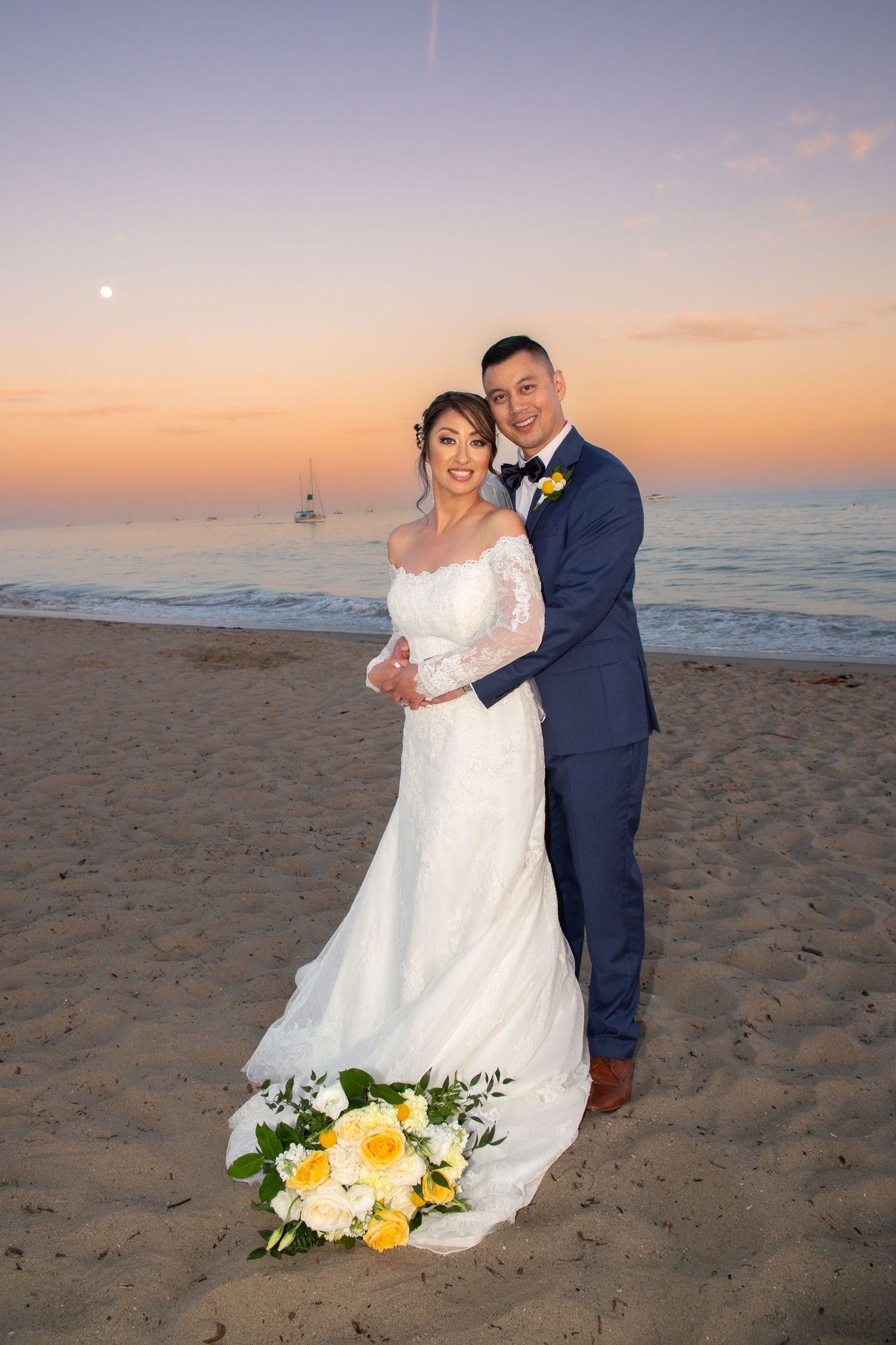 A couple in wedding attire embraces on a beach at sunset with yellow floral bouquet on the sand.