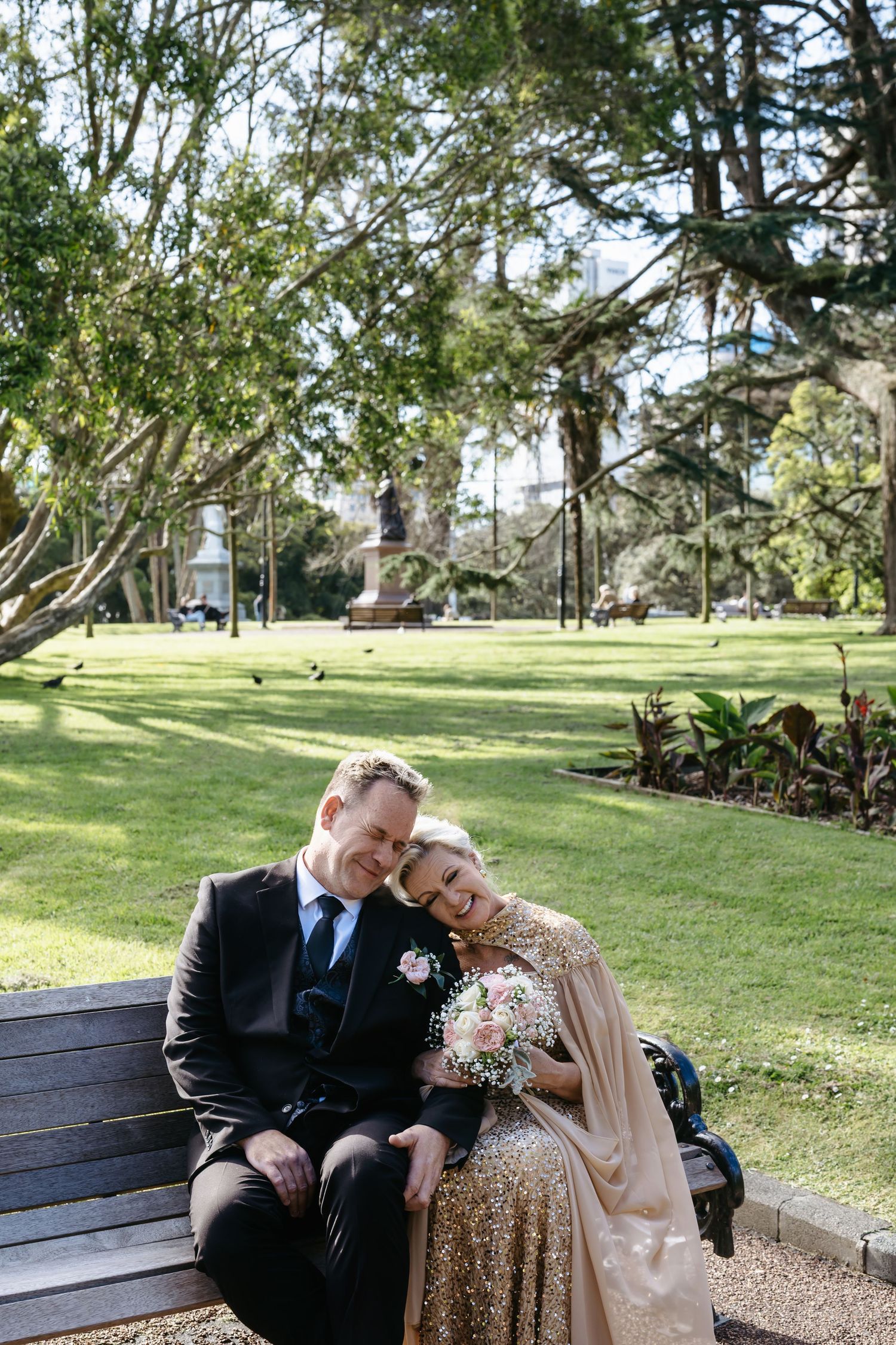 Bride in gold sequin dress and groom in black suit sit on park bench, sharing a tender moment with bridal bouquet.