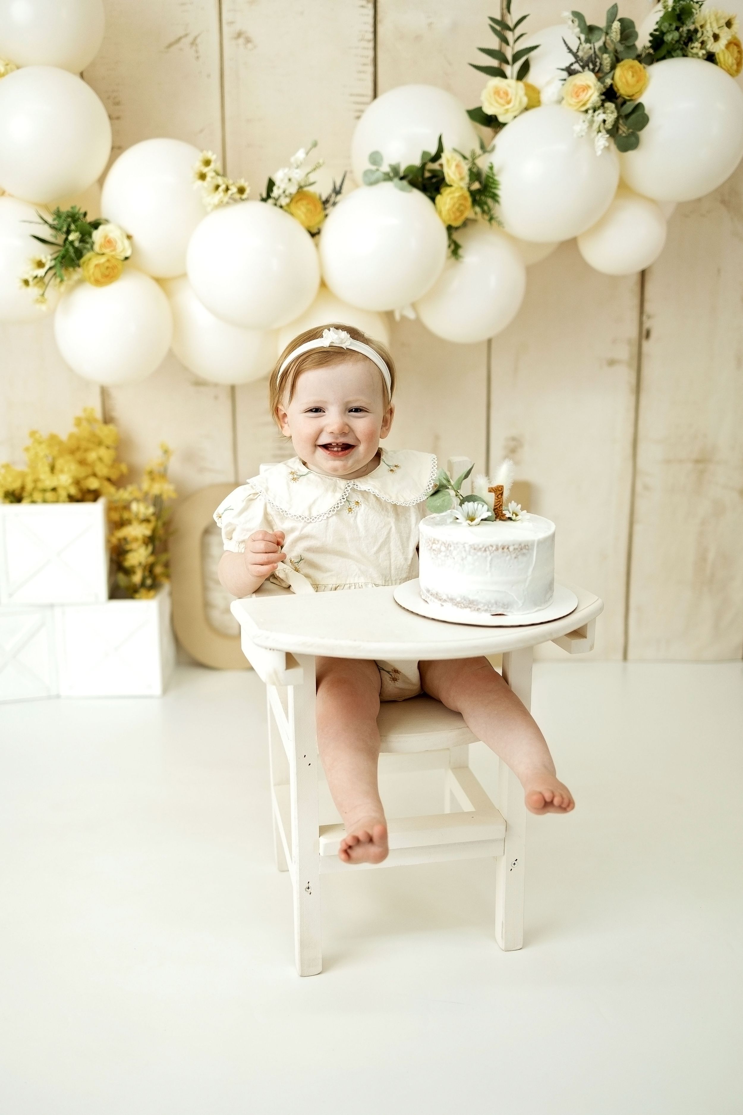 Smiling baby girl in white outfit sits in high chair with first birthday cake, surrounded by white and yellow balloon decorations.