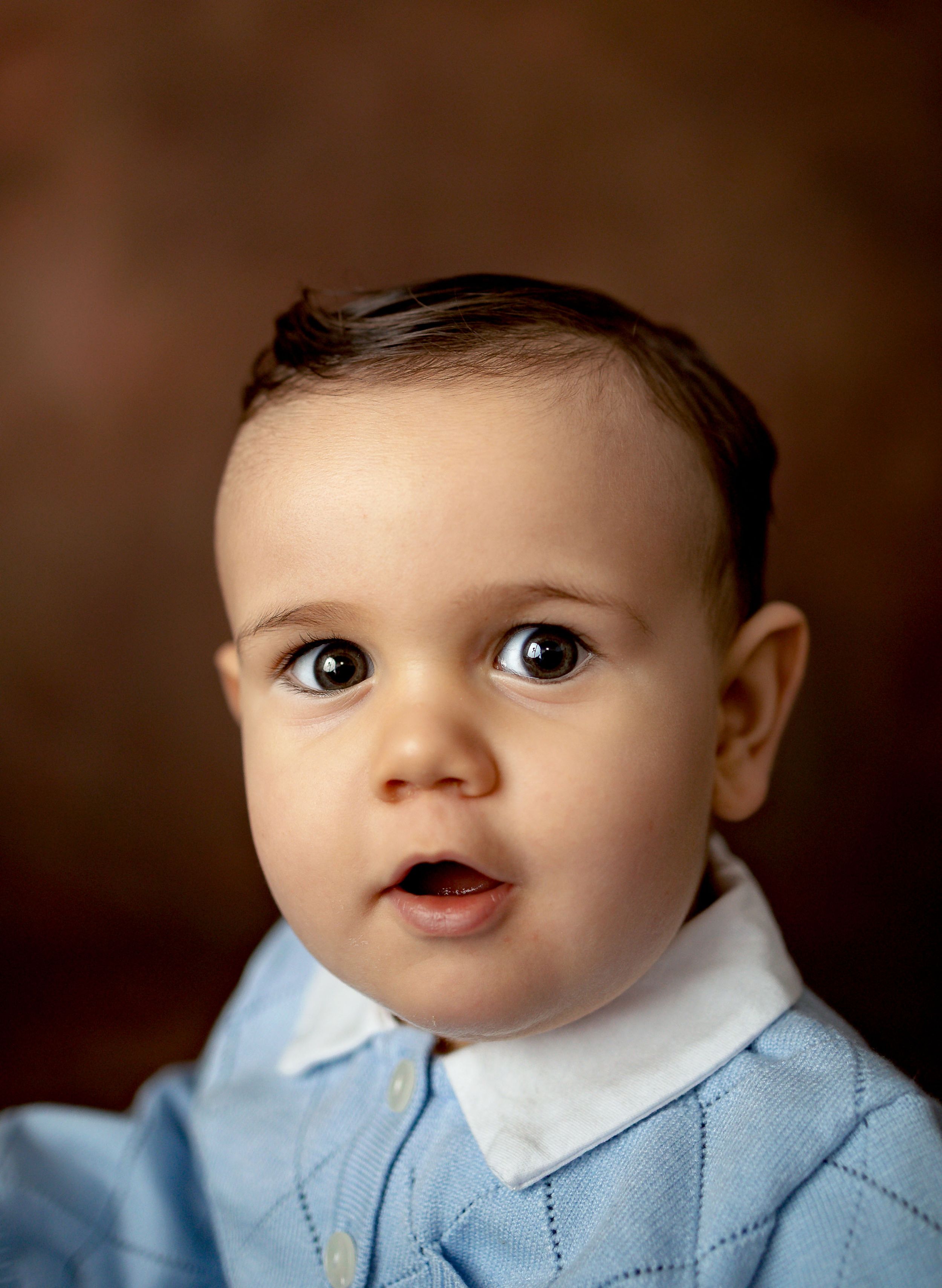 Adorable baby boy with dark eyes wearing a light blue knit sweater over a white collar shirt, portrait photo.
