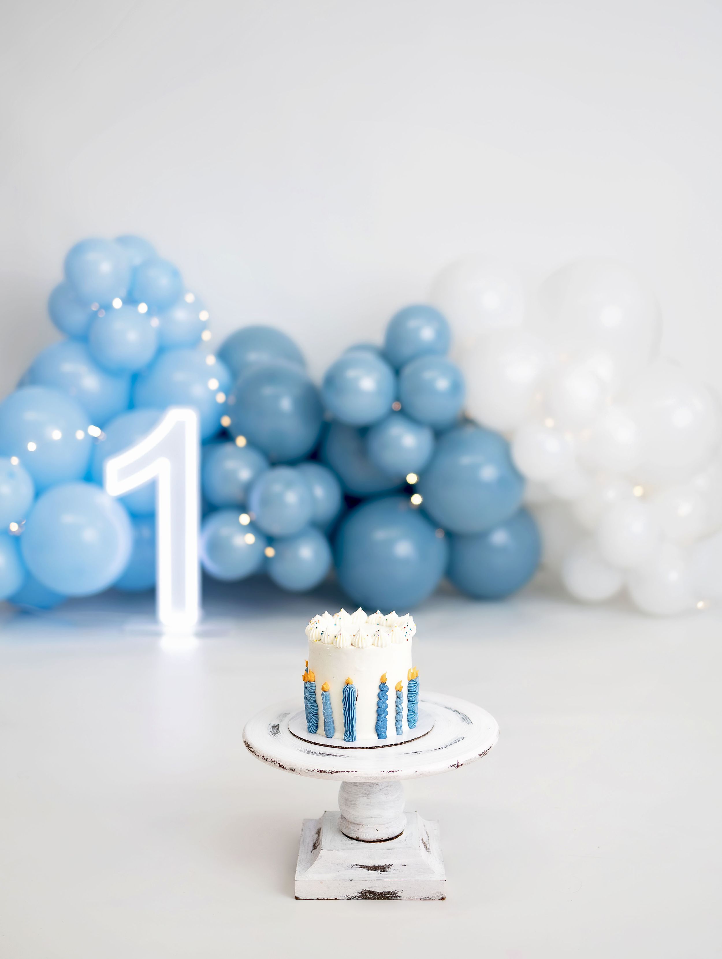 First birthday cake smash setup with blue and white balloon arch, number one decoration, and small decorated cake on white stand.