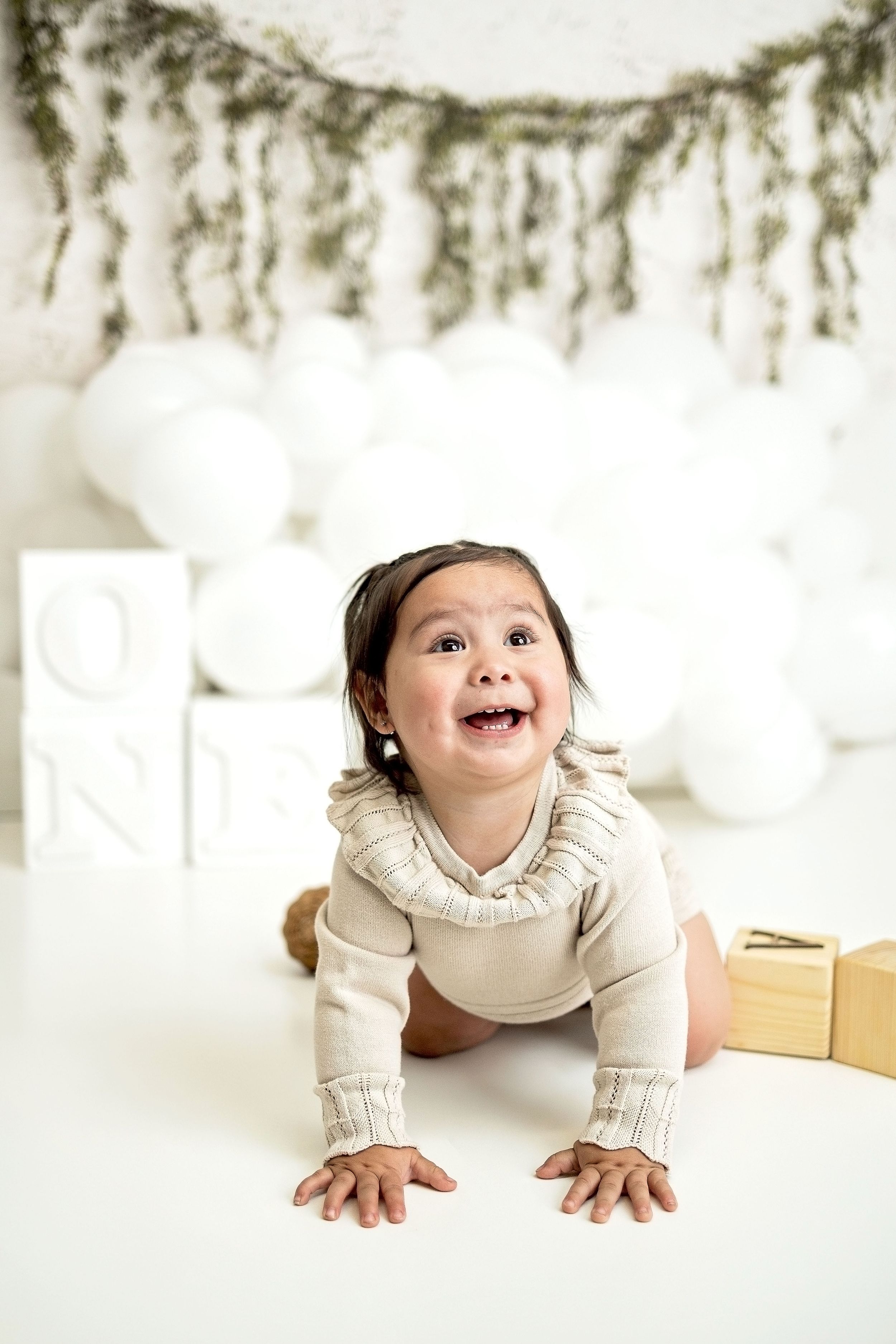 A happy baby girl in a beige onesie crawls on a white floor surrounded by balloons and letter blocks.