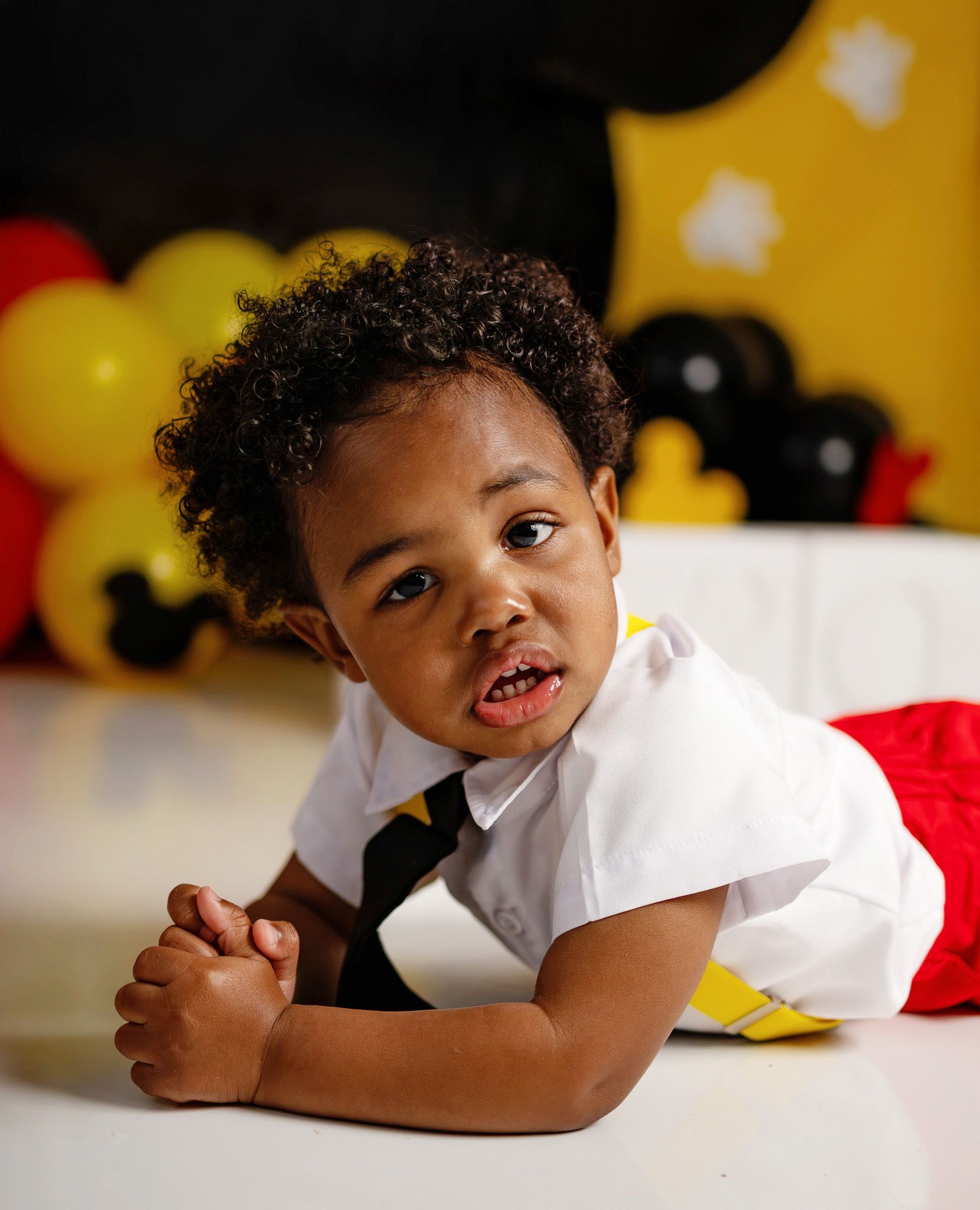 A young Black toddler boy in a white shirt and tie poses playfully at a Mickey Mouse themed birthday party.