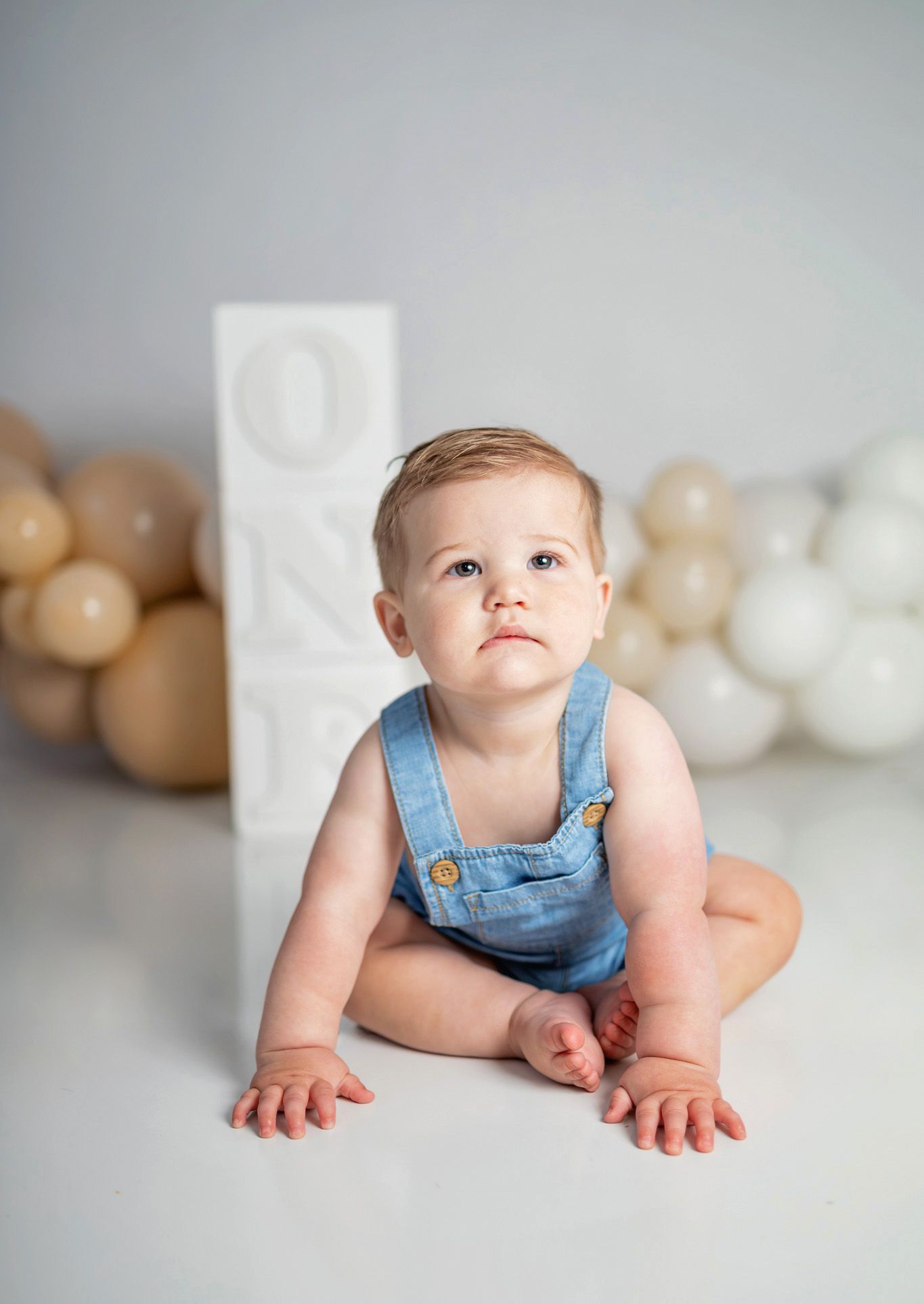 A baby in denim overalls sits on a white floor beside ONE blocks and beige balloons for a first birthday cake smash photo.