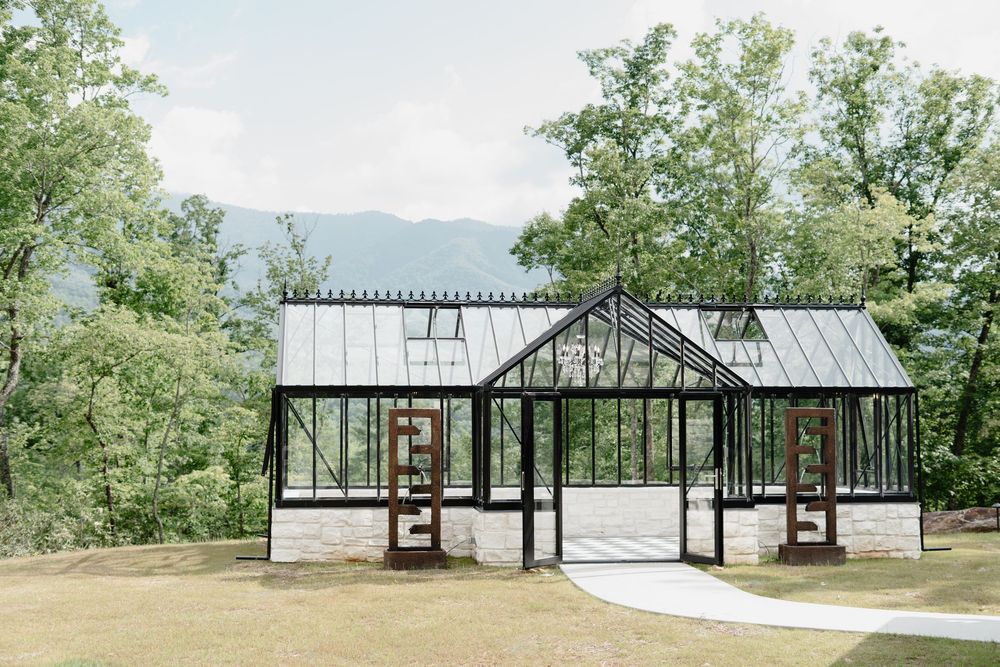 A stunning glass greenhouse wedding venue with black frames and stone base, surrounded by lush trees and mountain views.