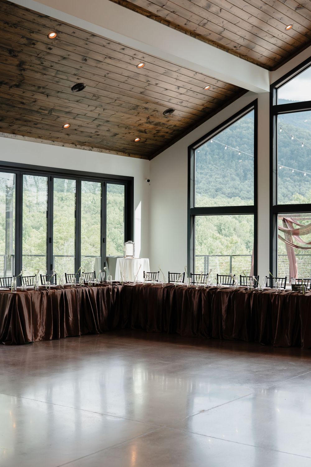 Elegant wedding reception hall with brown satin head table, chiavari chairs, wood ceiling, and floor-to-ceiling windows with mountain views.