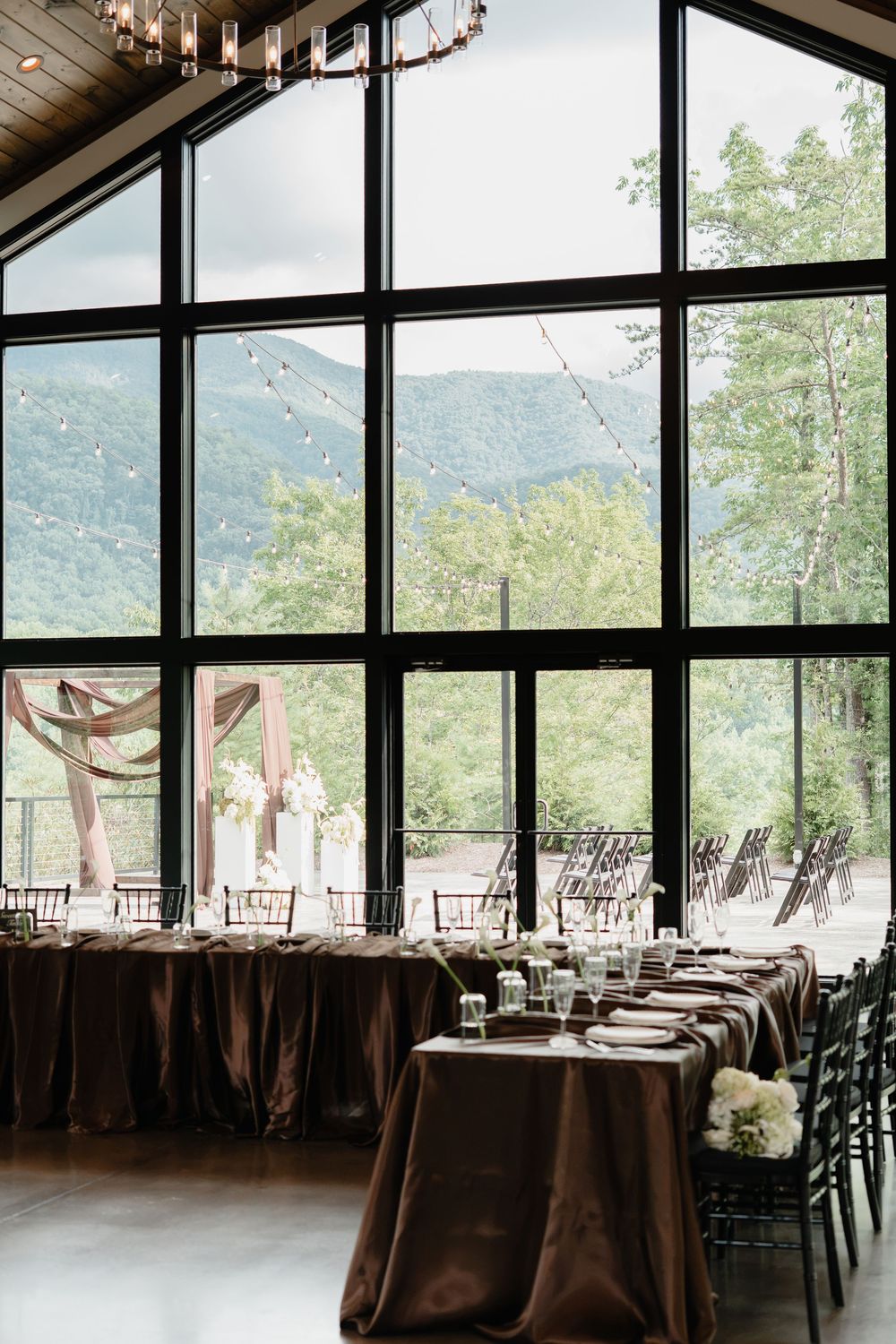 Elegant wedding reception hall with floor-to-ceiling windows, mountain views, brown satin tablecloths, and string lights.