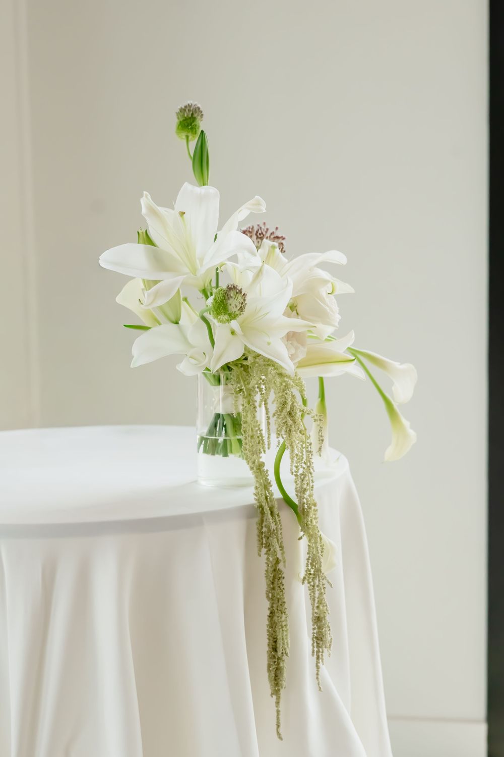 Elegant white lily and greenery floral arrangement in a glass vase on a white draped table.