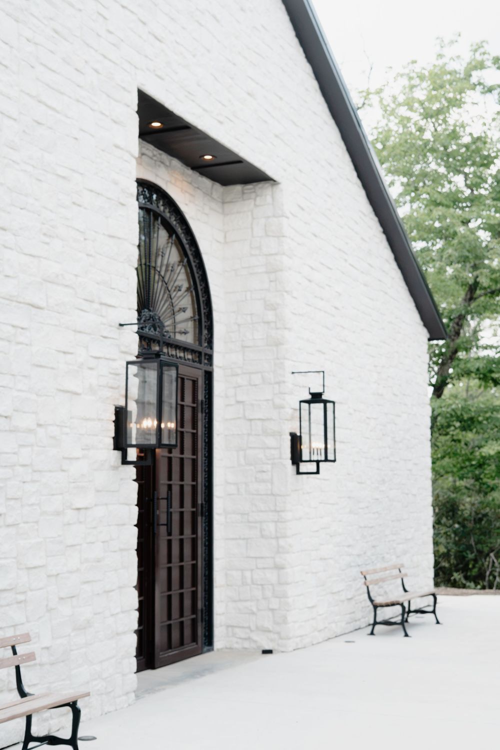 White brick wedding venue exterior featuring an arched iron door, black lanterns, and wooden benches.