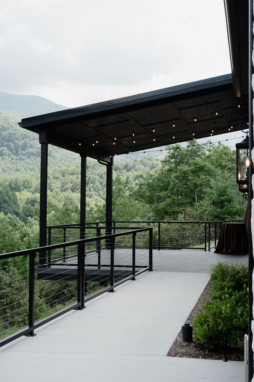 Modern covered patio with string lights overlooking lush mountain forest views, featuring sleek black metal railings and columns.