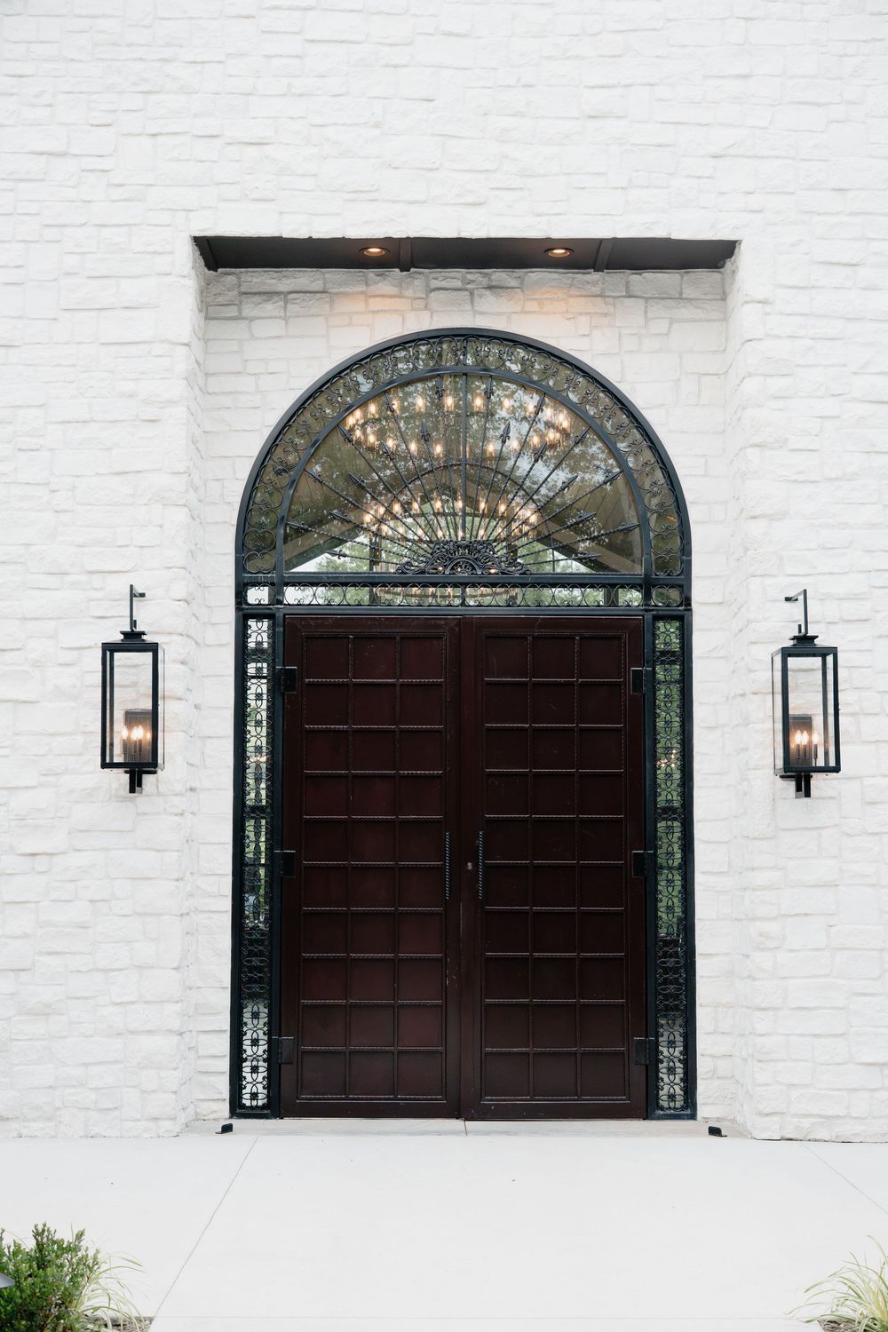 Elegant arched double doors with dark wood panels, ornate iron details, and wall lanterns on a white brick facade.
