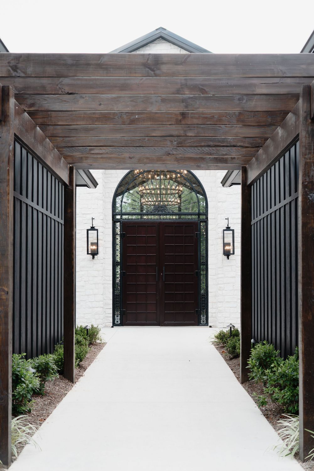 Grand venue entrance with dark wood pergola, black metal gates, white brick walls, and arched double doors with chandelier.