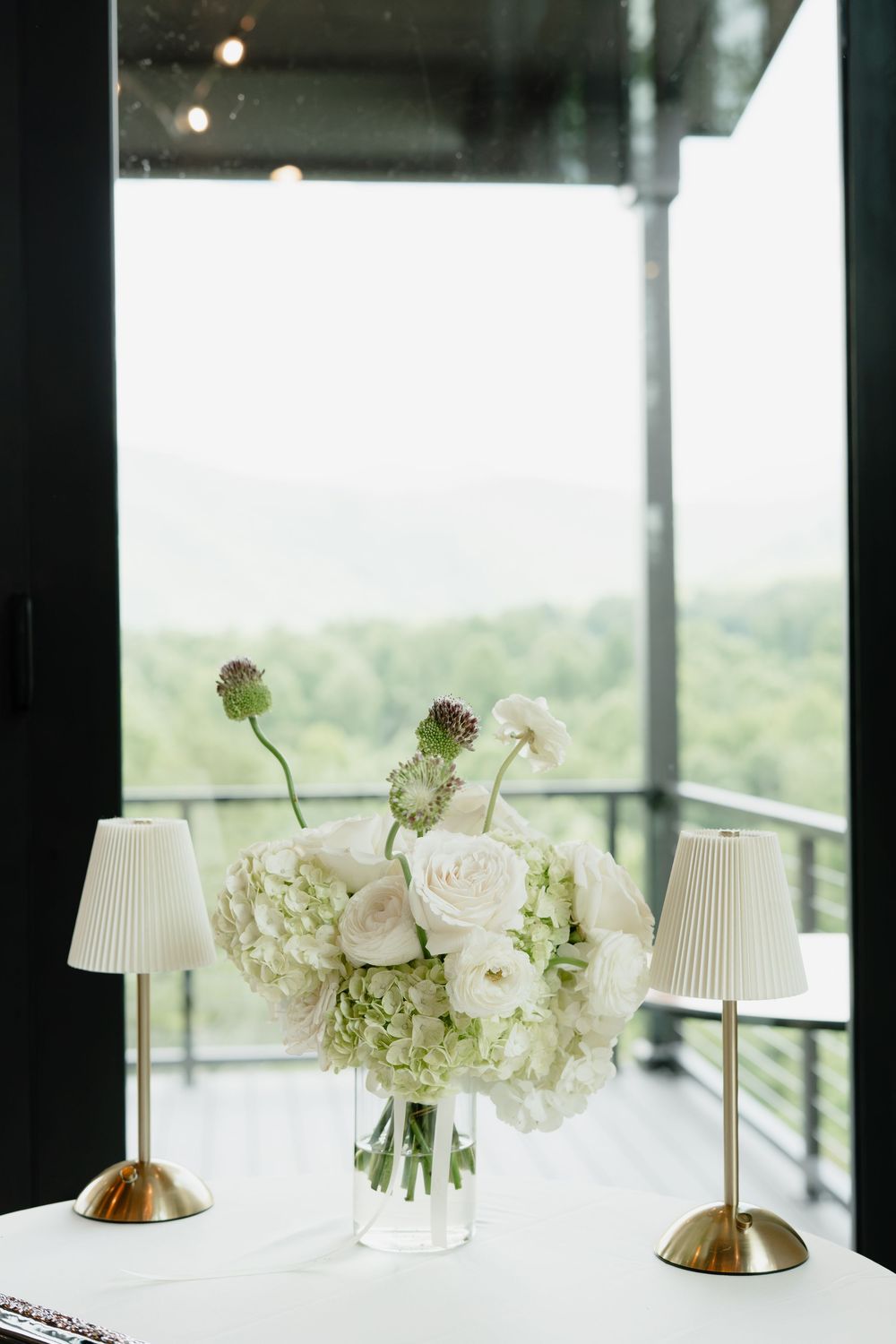 White floral centerpiece with hydrangeas and roses in glass vase flanked by gold table lamps against panoramic window.