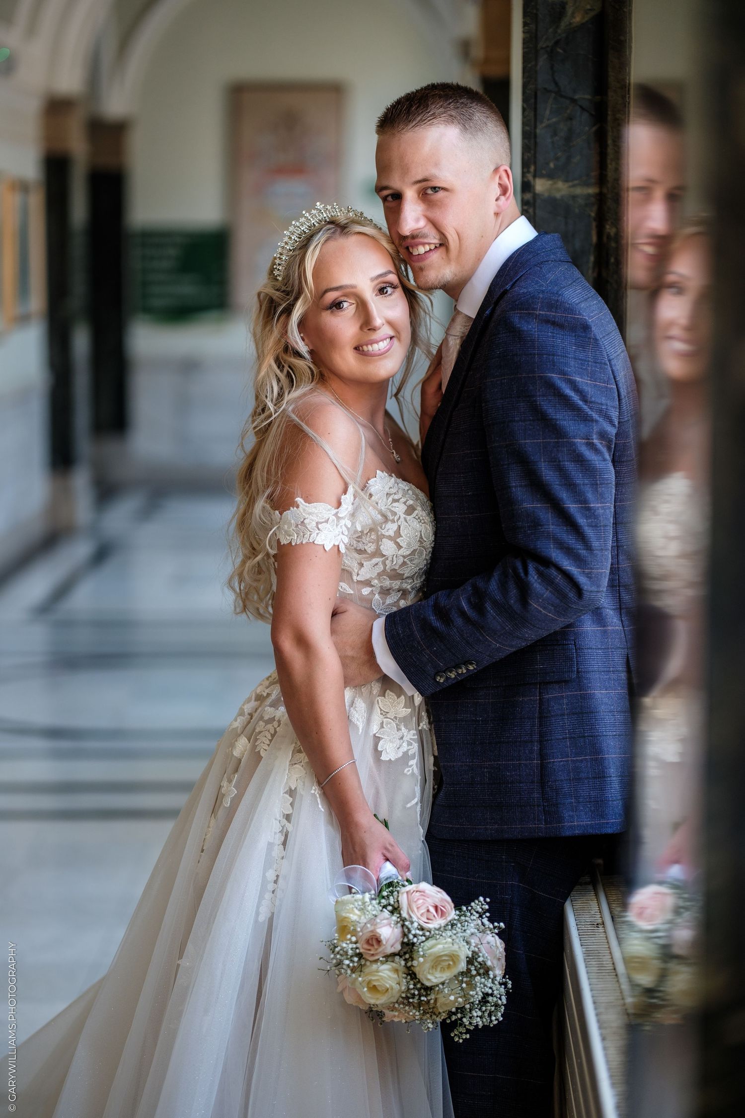 Bride in lace gown holding bouquet poses with groom in blue suit, their reflection visible in a mirror behind them.
