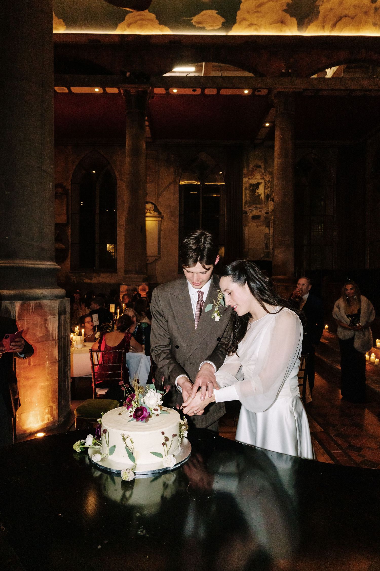 Bride and groom cutting white floral wedding cake together in elegant candlelit venue with stone columns.