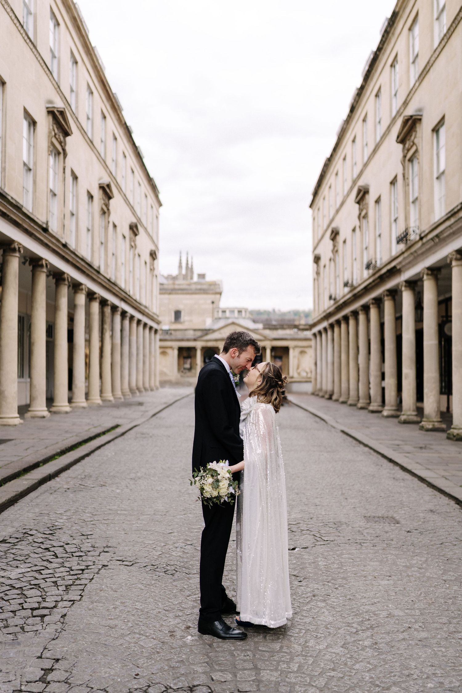 Bride and groom share a romantic moment in a grand colonnaded street, bride holding a white bouquet in an elegant wedding dress.