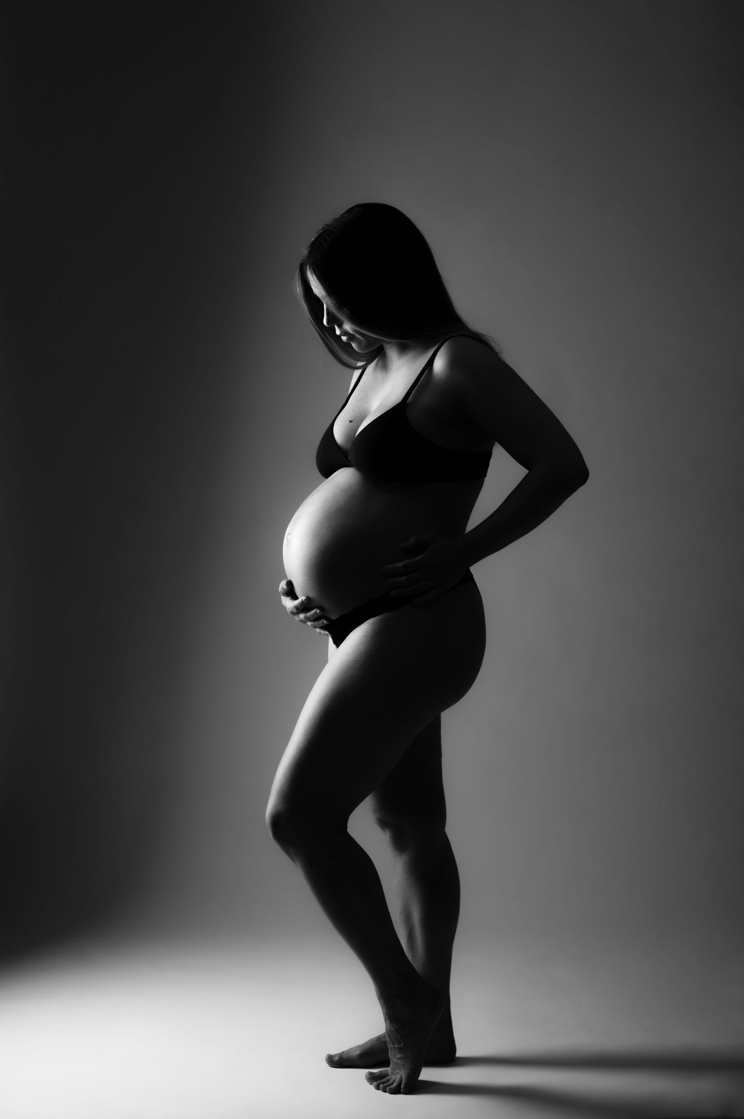 Black and white maternity photo of a pregnant woman in lingerie cradling her baby bump in a studio.