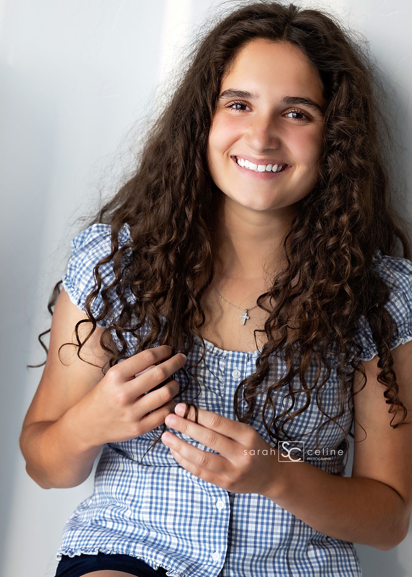 Smiling teenage girl with long curly brown hair wearing a blue gingham top, posing for a professional portrait photo.