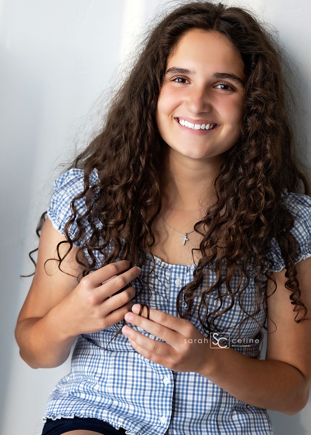 Smiling teenage girl with long curly brown hair wearing a blue gingham top, posing for a professional portrait photo.