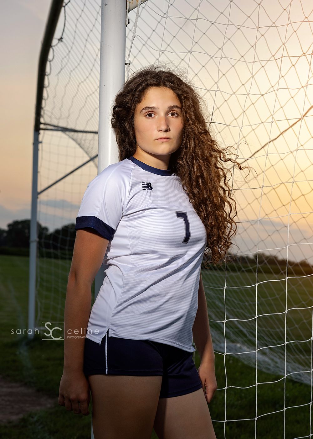 Female soccer player #7 in white and navy uniform poses by goal net at sunset on a green field.