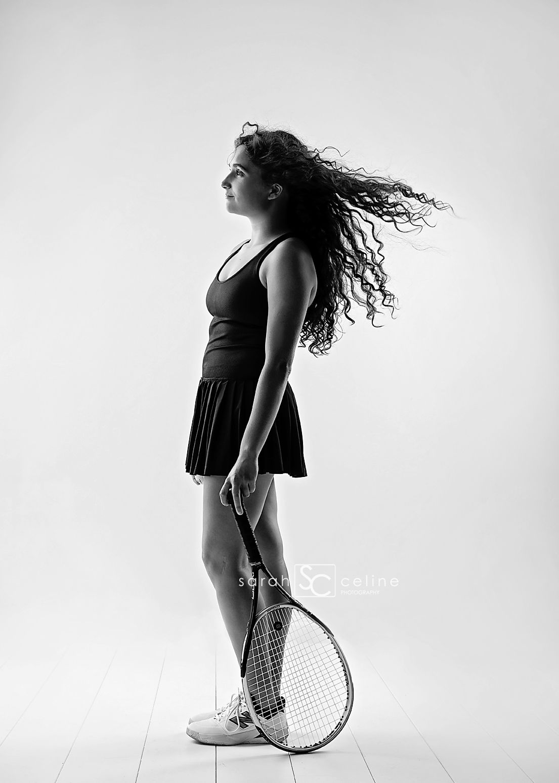 Black and white photo of a young woman in tennis attire holding a racket, her curly hair flowing dramatically in the wind.