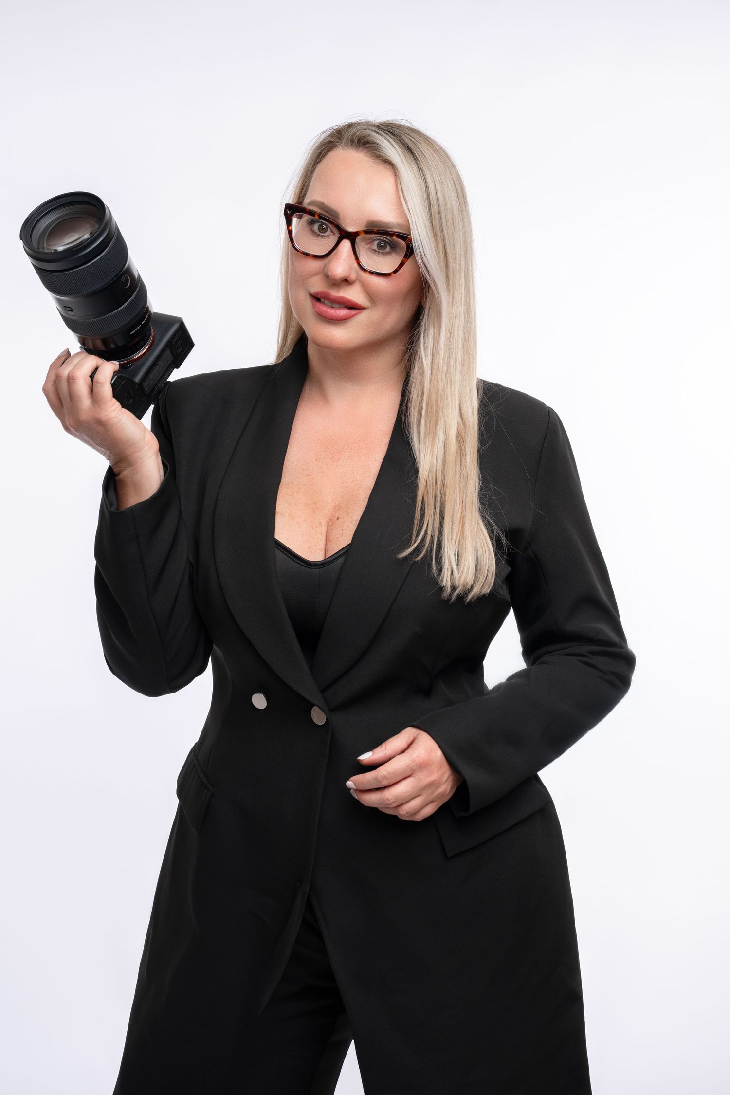 Professional female photographer in black suit holding DSLR camera with telephoto lens, wearing glasses, white background.