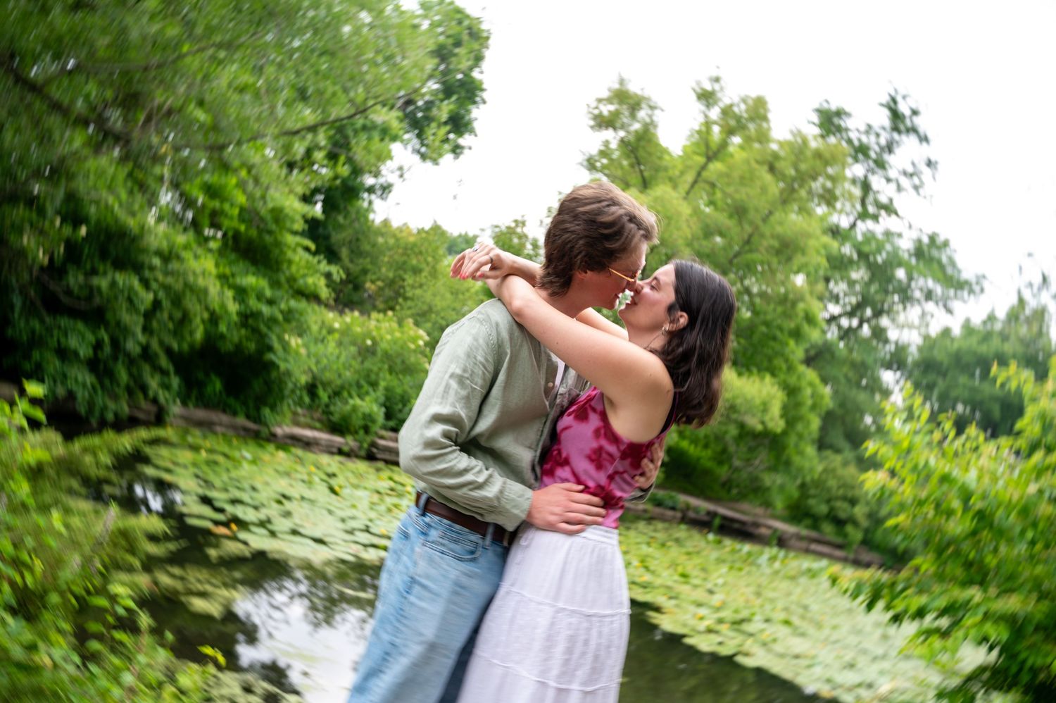 A couple shares a romantic kiss near a serene lily pad pond surrounded by lush green trees outdoors.