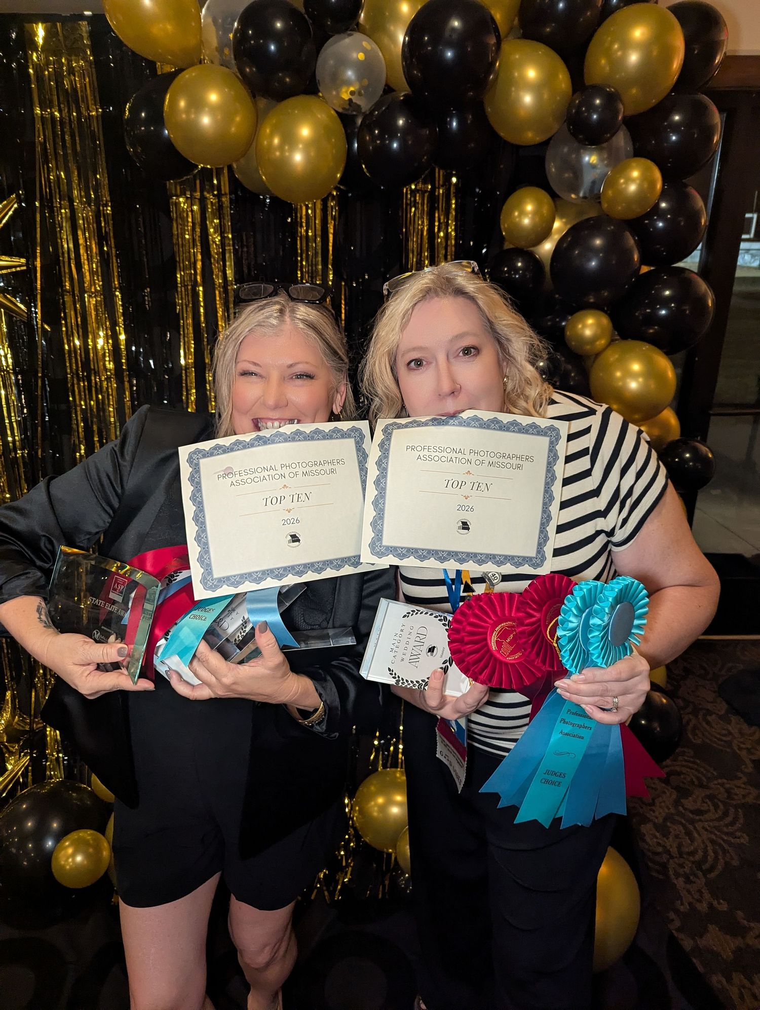Two women proudly display Top Ten awards, certificates, and ribbons at a photography competition with gold and black balloon decor.