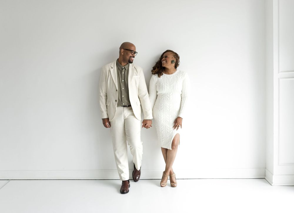 A couple standing against white wall smiling at each other during an engagement session at a natural light studio.