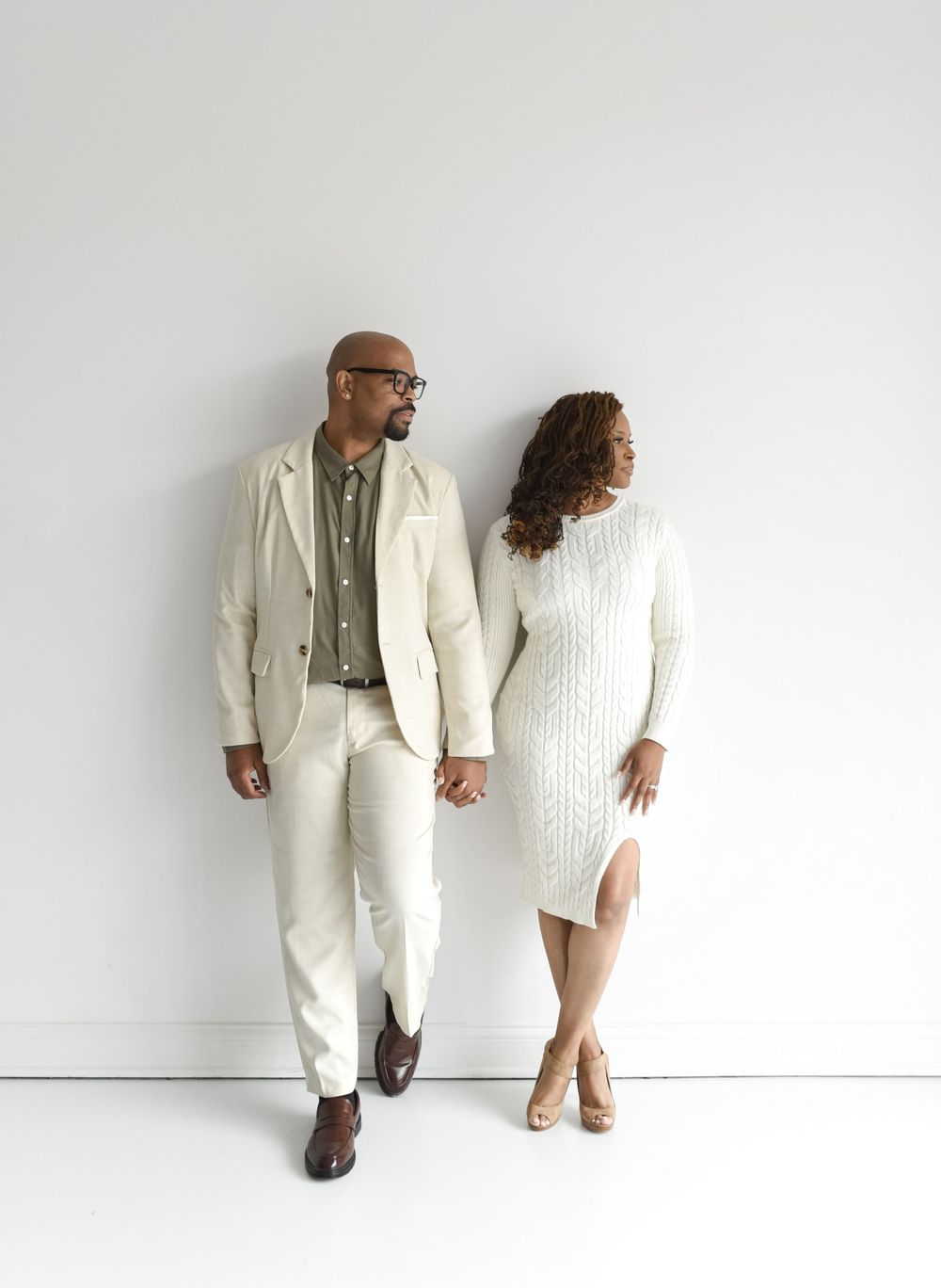 A couple standing against white wall looking towards window during an engagement session at a natural light studio.