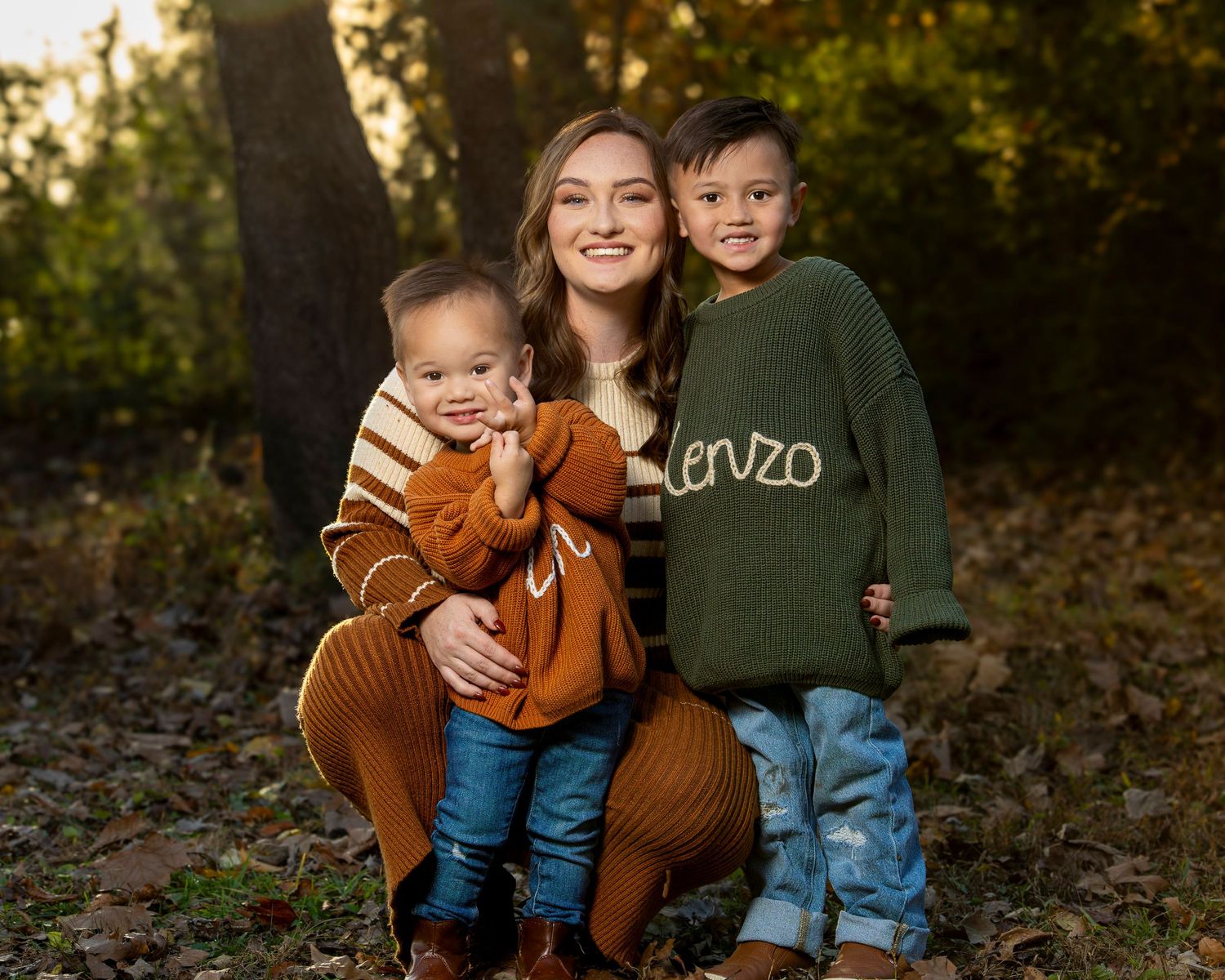A smiling young woman crouches outdoors with two young boys in autumn woods, wearing colorful sweaters in warm fall tones.