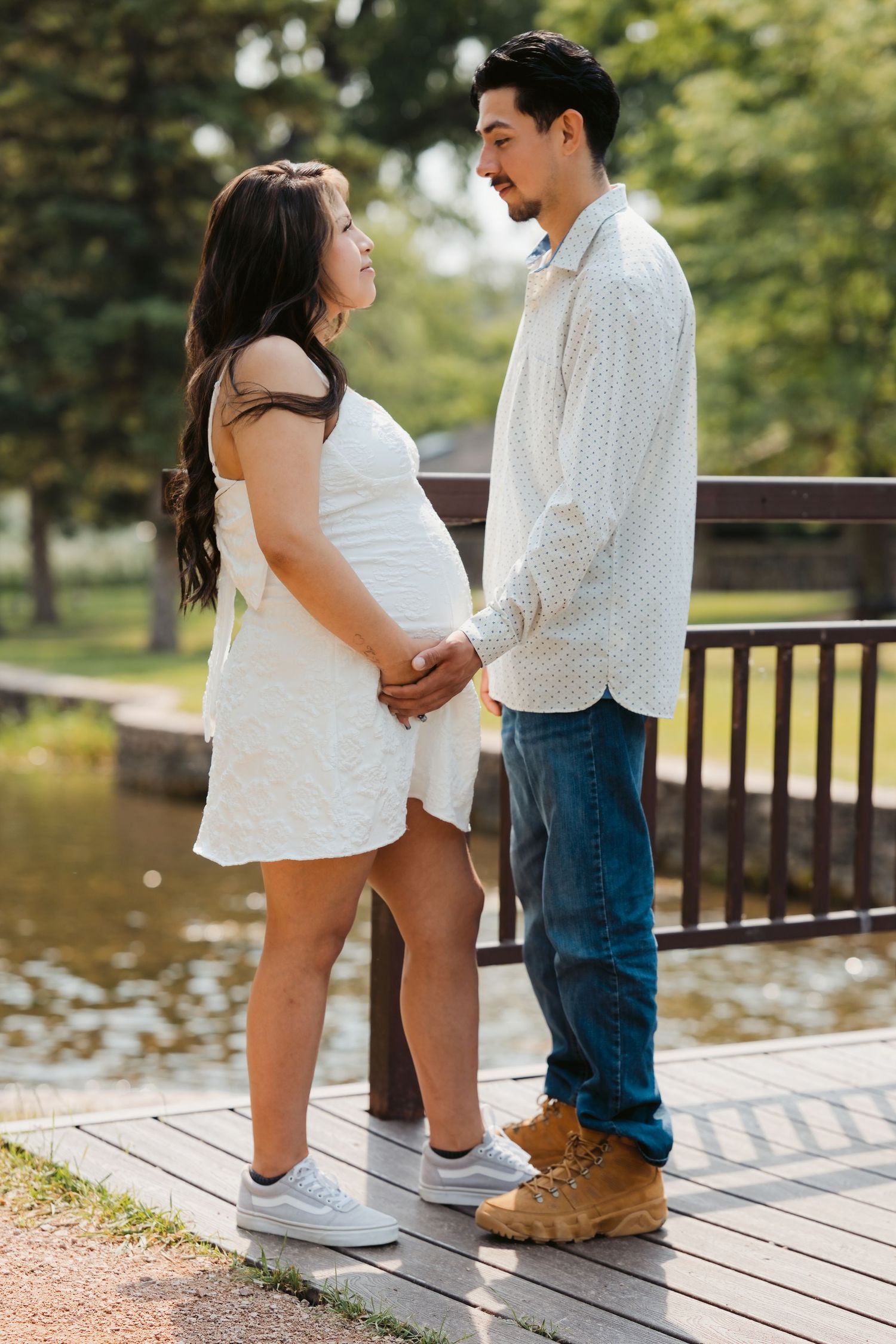 Pregnant couple holding hands on a wooden dock in a park, woman in white dress, man in jeans and boots.