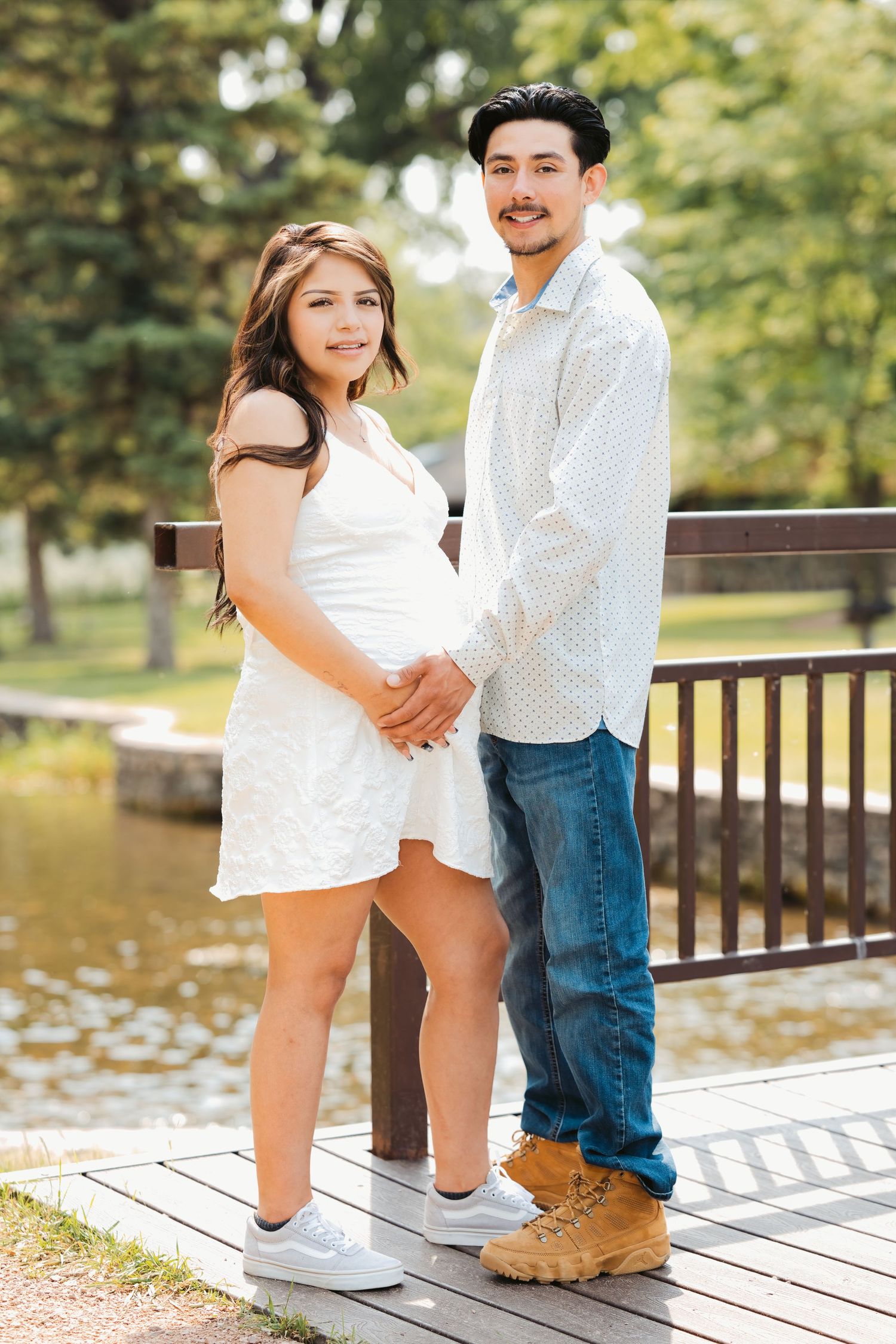 A young couple holds hands on a wooden bridge in a park, the woman in a white dress cradling her baby bump.