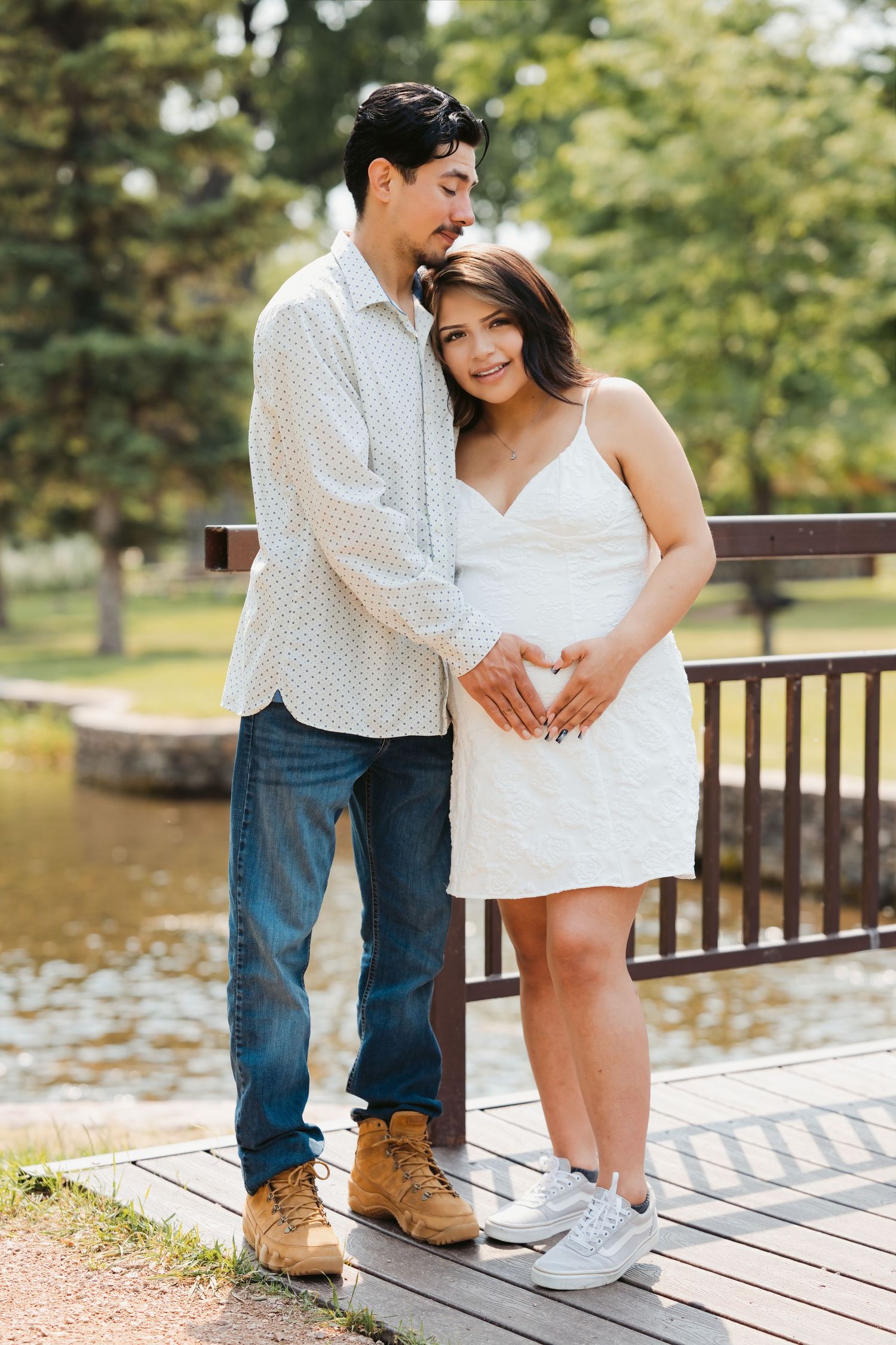 Couple on wooden dock, man embracing pregnant woman from behind, both forming heart shape over baby bump in park.