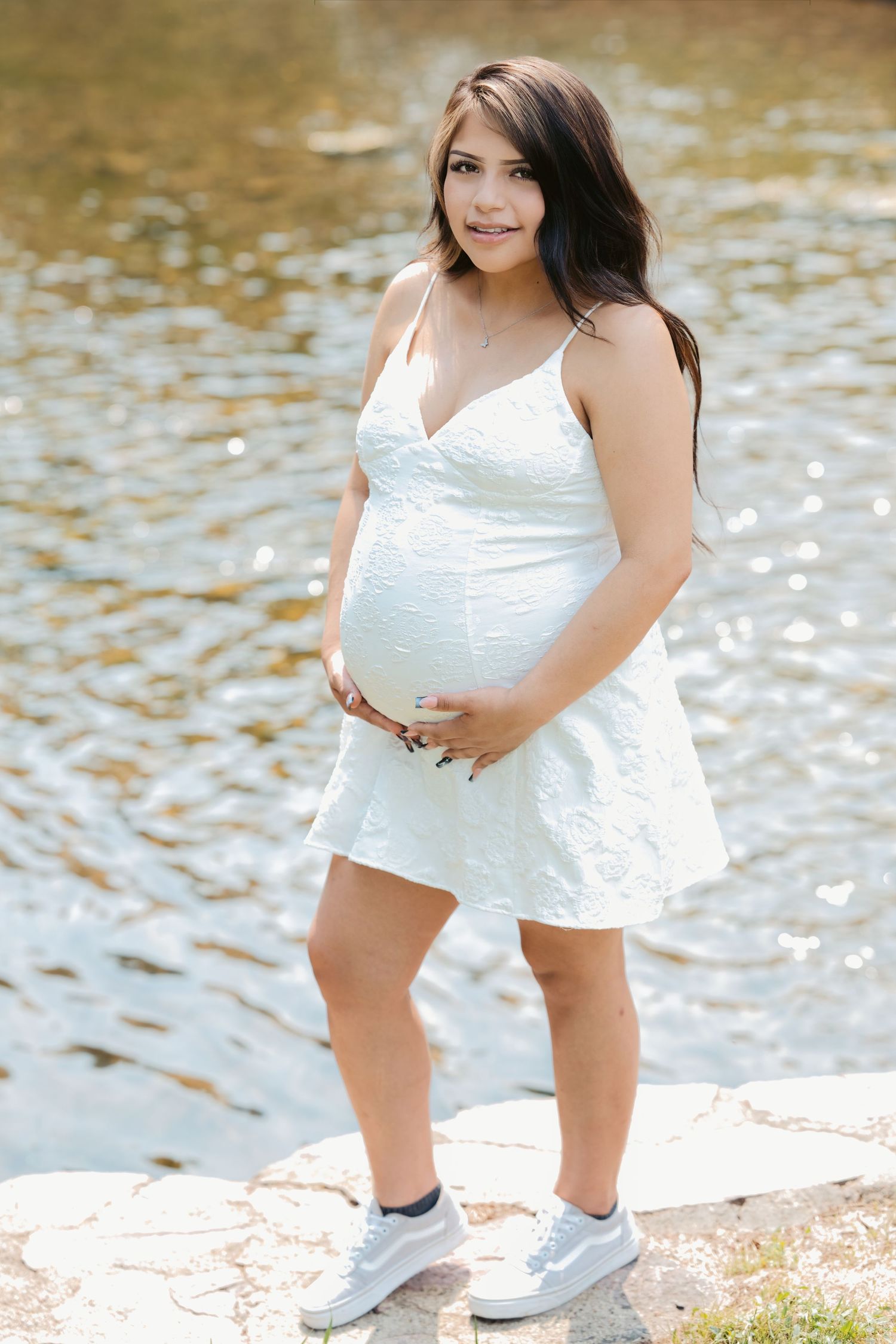 Pregnant woman in white sundress holding baby bump, standing by a sparkling lake on a sunny day.