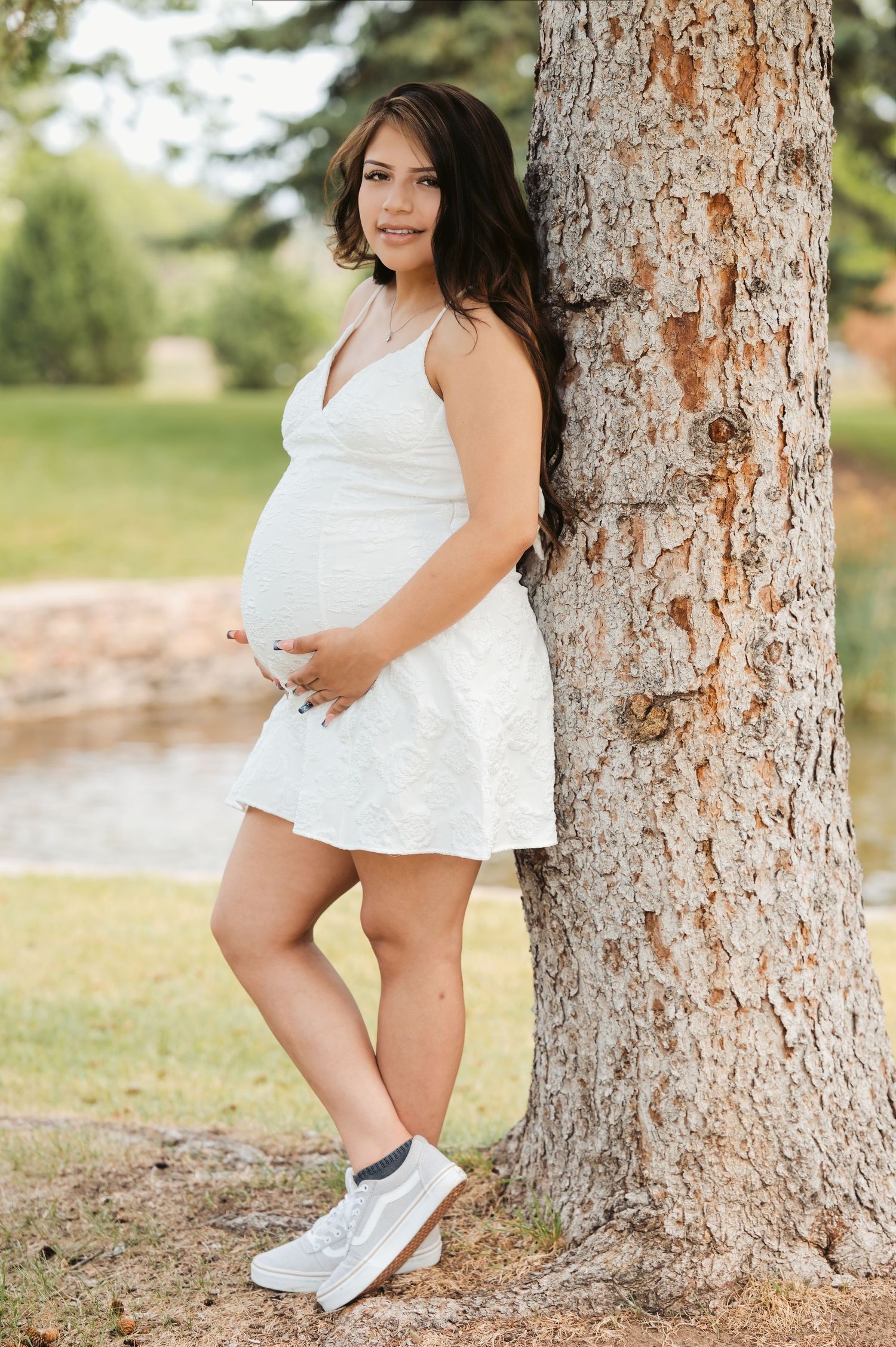 Pregnant woman in white dress leaning against tree in park, cradling baby bump, wearing white sneakers outdoors.