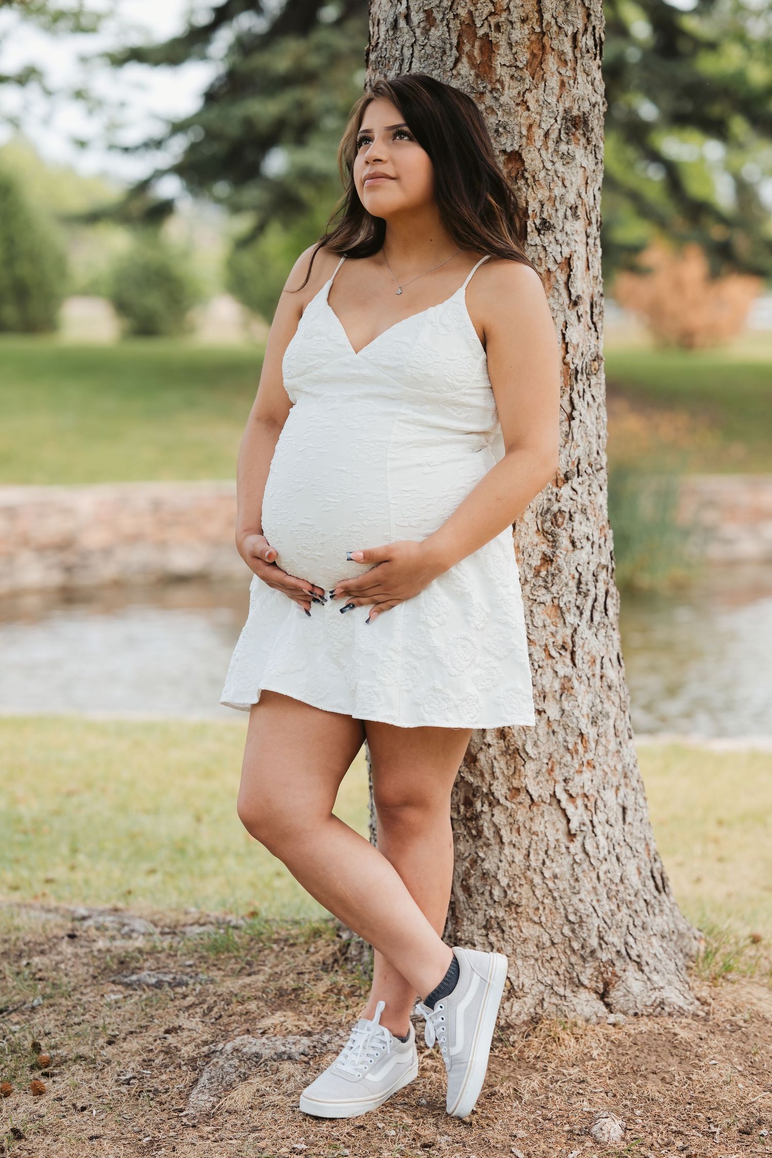 Pregnant woman in white sundress and sneakers leaning against tree in park, cradling baby bump.