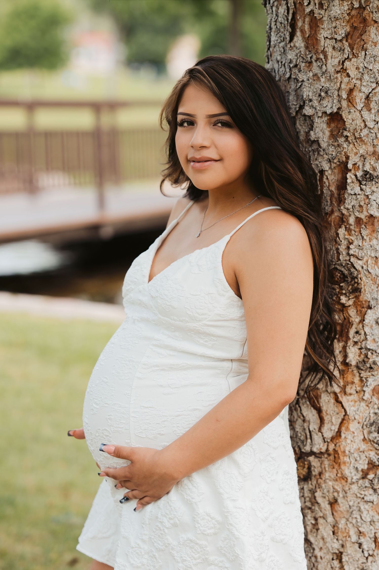 Pregnant woman in white dress leaning against tree trunk outdoors, cradling baby bump, with bridge and greenery in background.