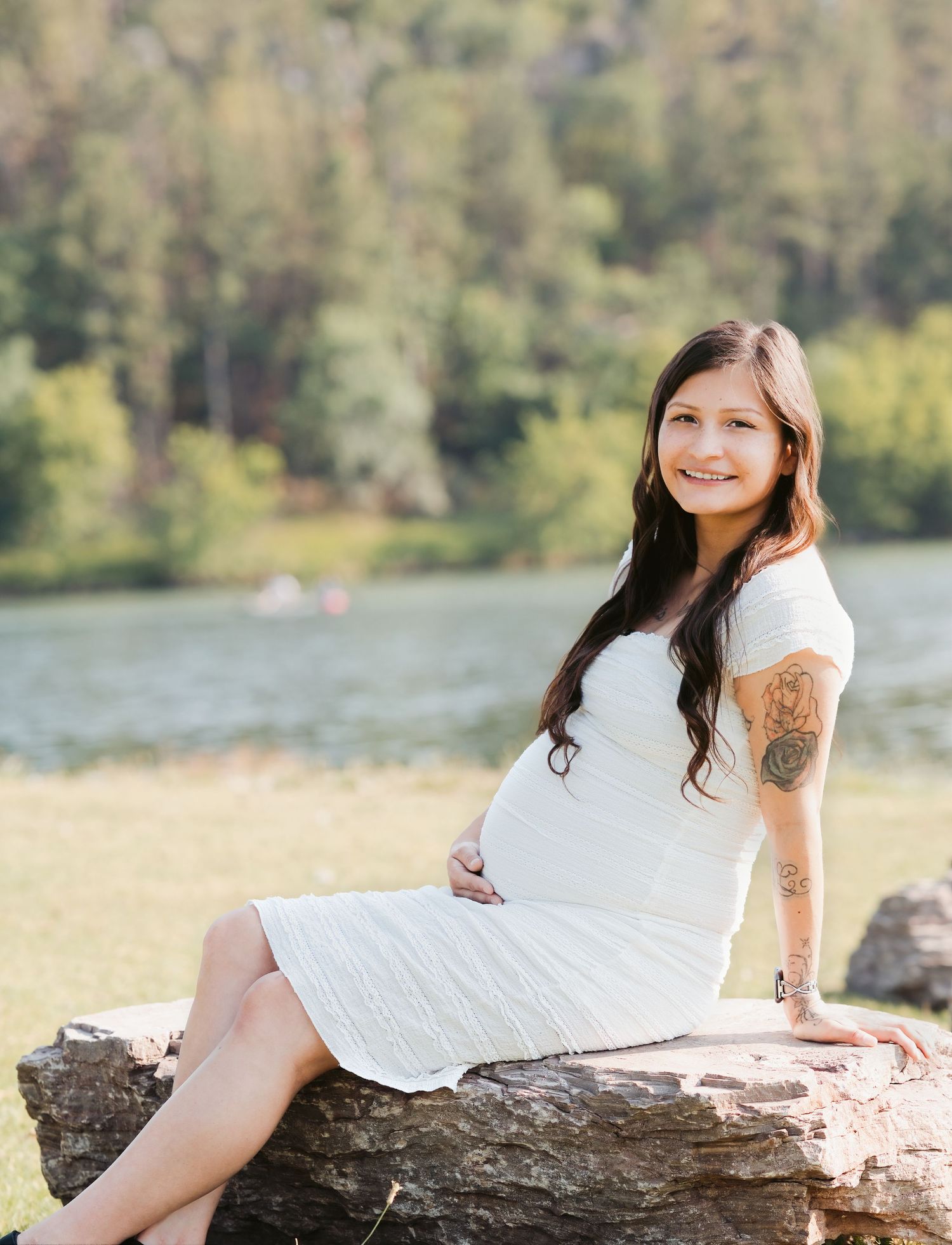Smiling pregnant woman in white dress sitting on rocks outdoors near a lake, showing baby bump and arm tattoo.